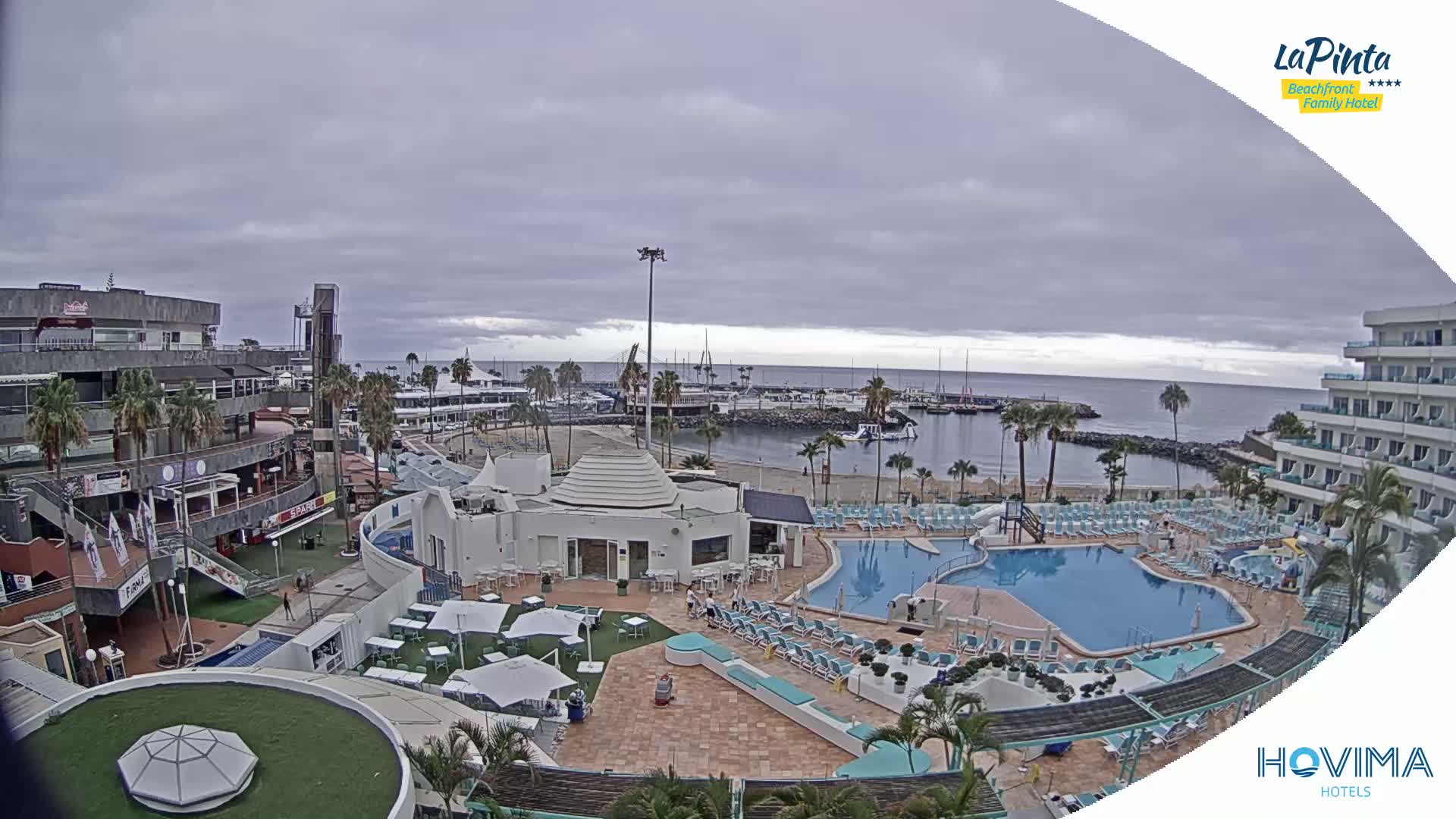 A resort with swimming pools and a beach is shown under an overcast sky, overlooking a marina with boats.