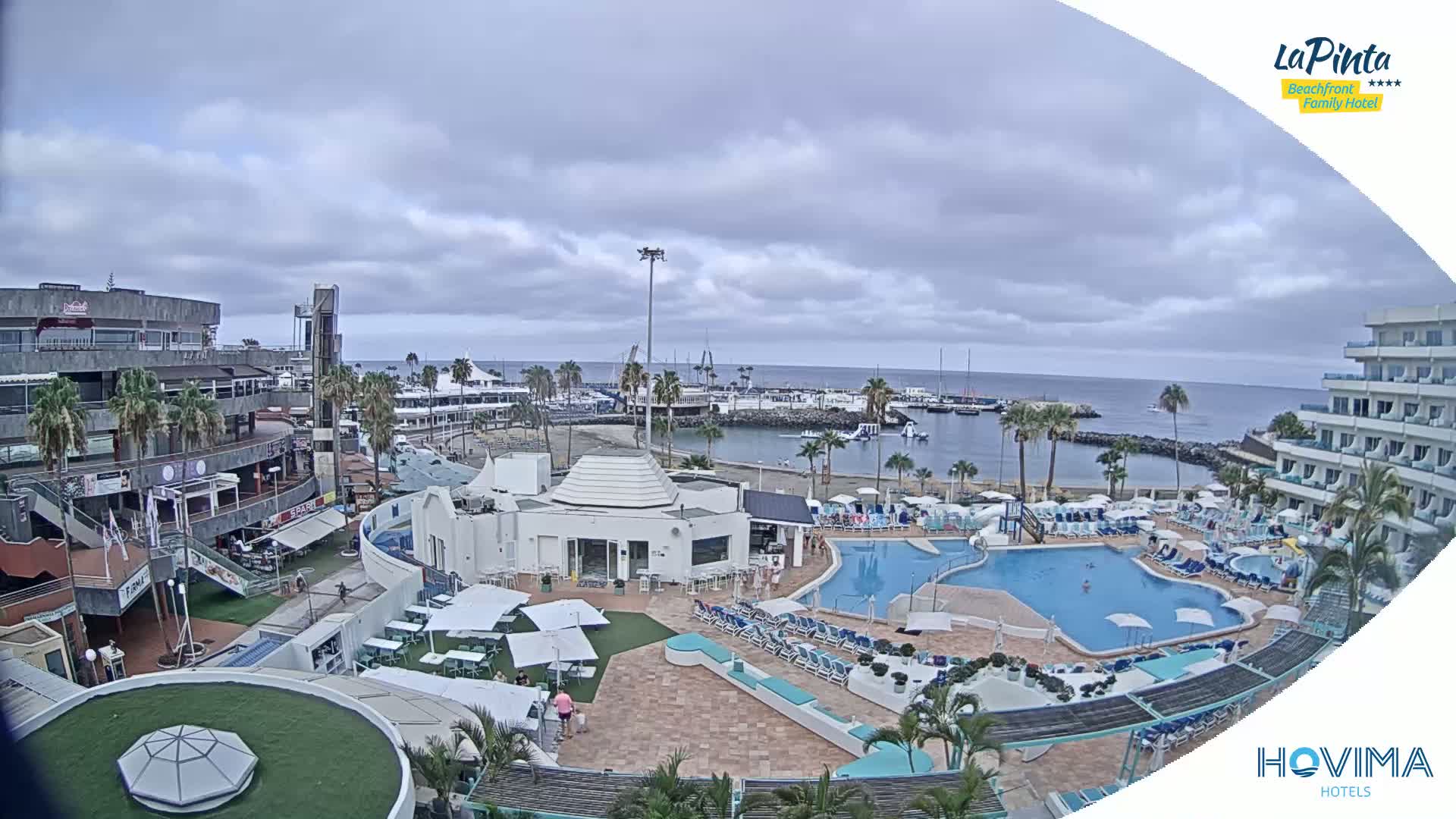A resort with swimming pools overlooks a marina and calm ocean under a cloudy sky.