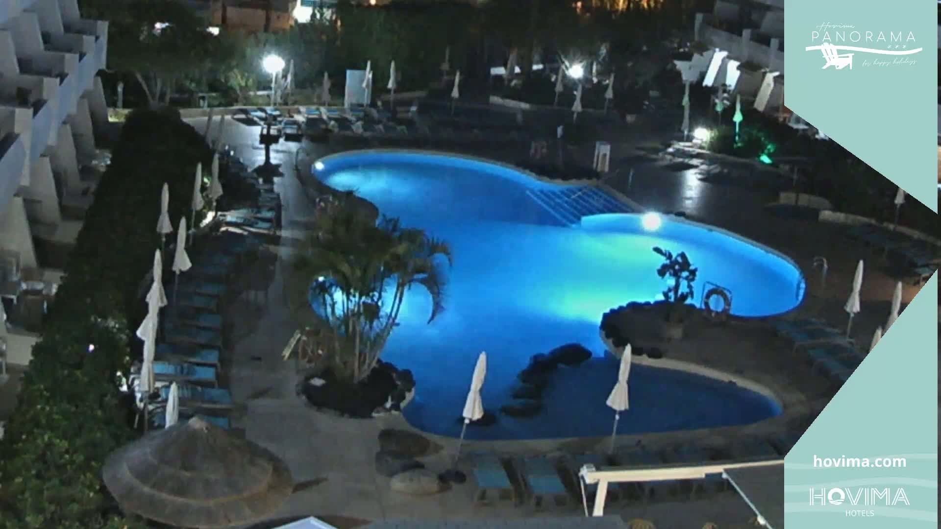 A brightly lit, kidney-shaped swimming pool is surrounded by lounge chairs and umbrellas at night.