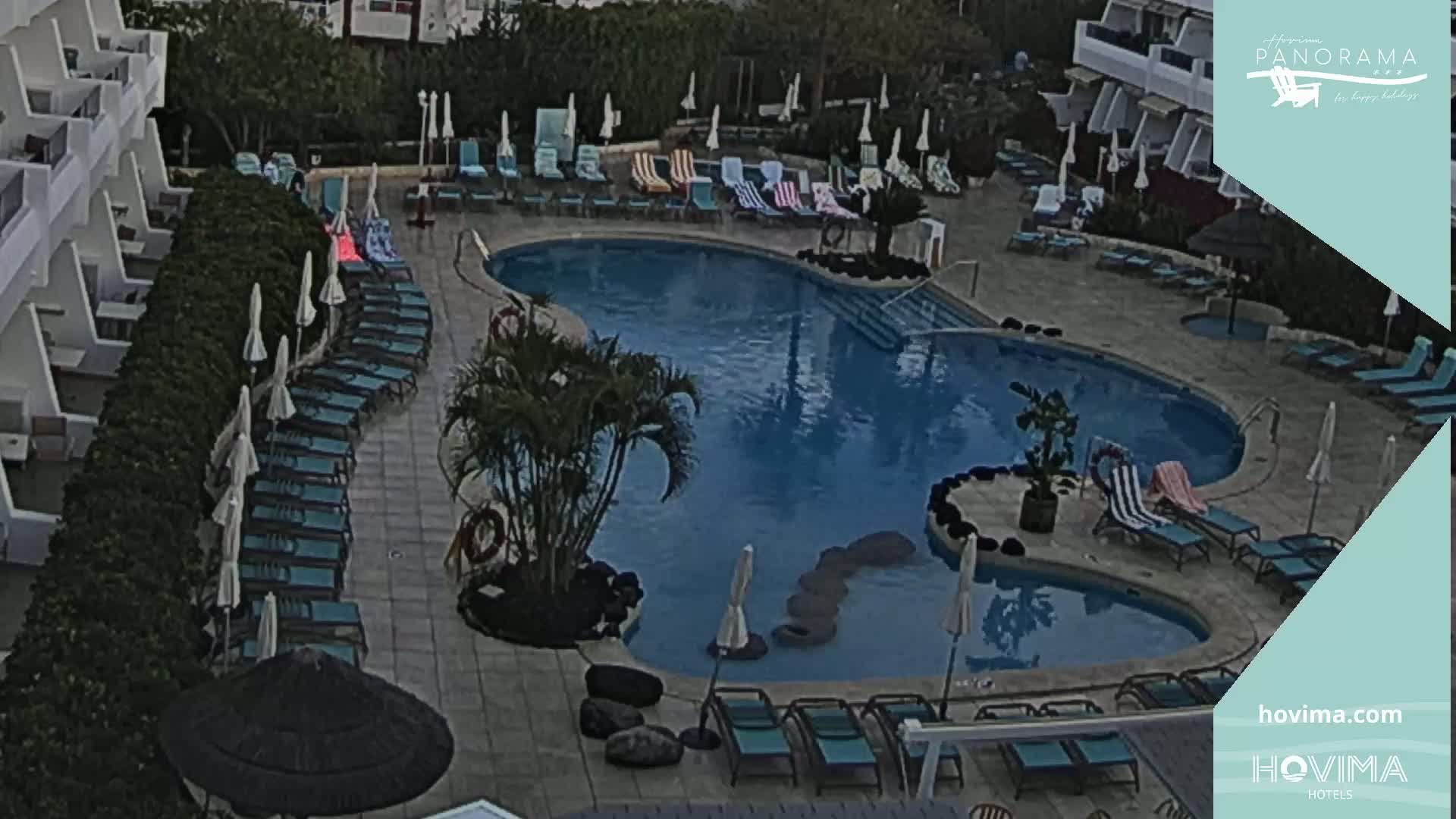 An outdoor swimming pool surrounded by lounge chairs and umbrellas, situated near a building, under an overcast sky.