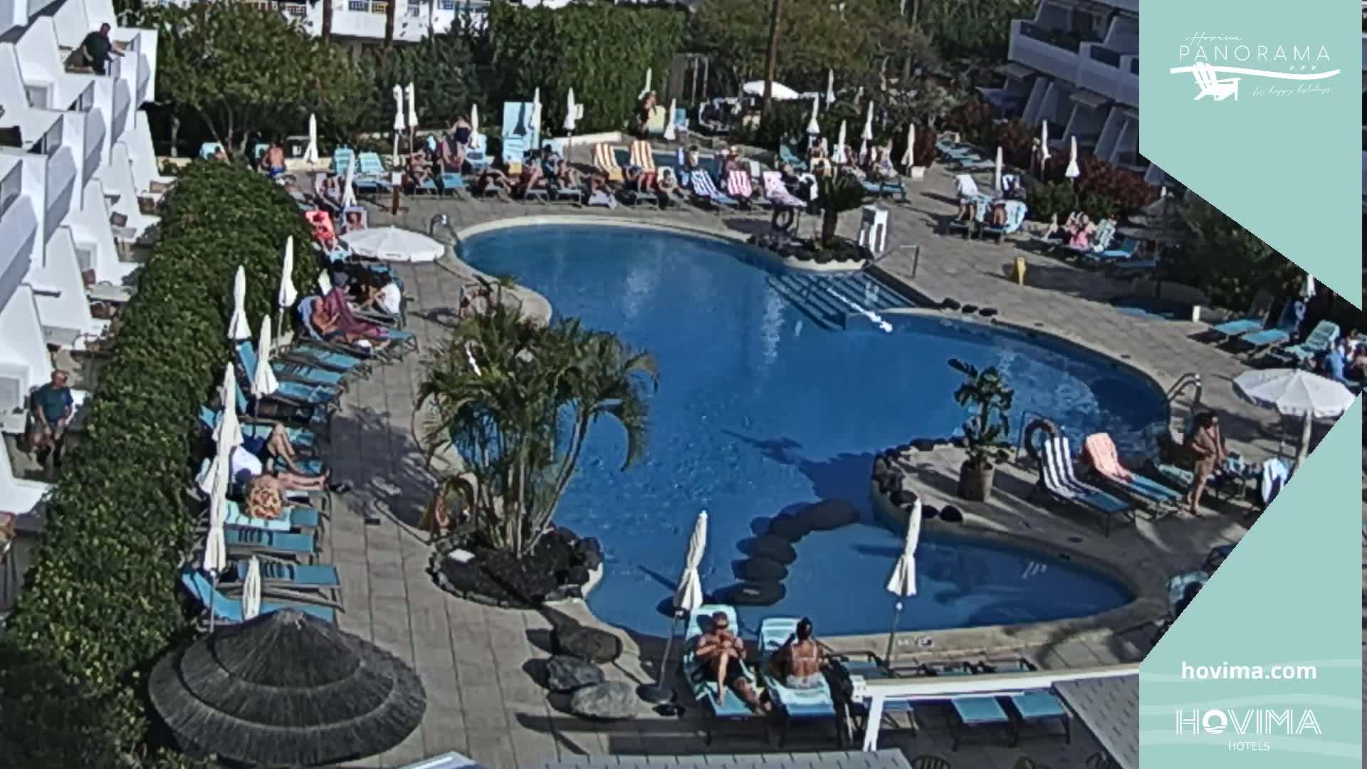 A sunny outdoor scene shows a kidney-shaped swimming pool surrounded by lounge chairs and umbrellas, with many people relaxing around the pool area.