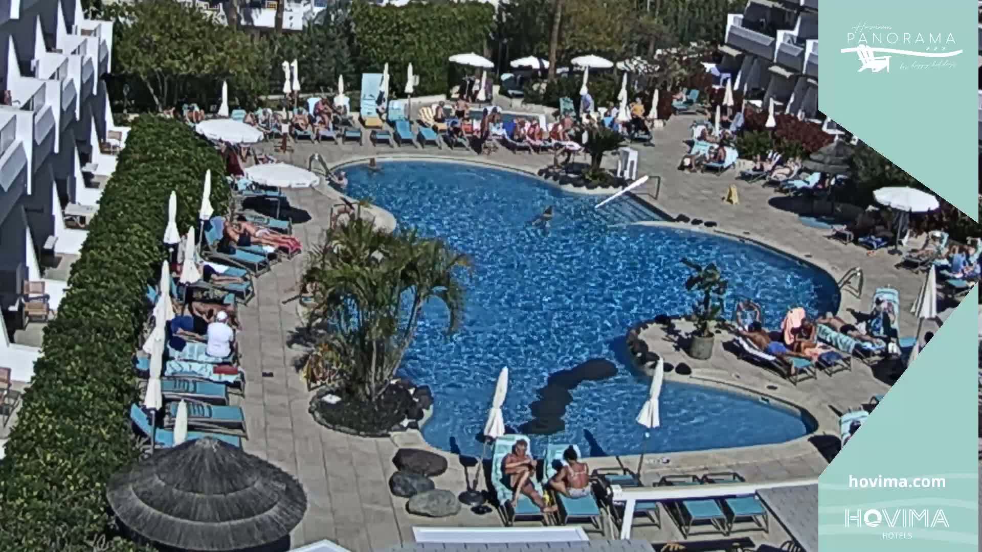 A sunny outdoor scene shows a kidney-shaped swimming pool surrounded by people lounging on chairs and umbrellas near a white building.