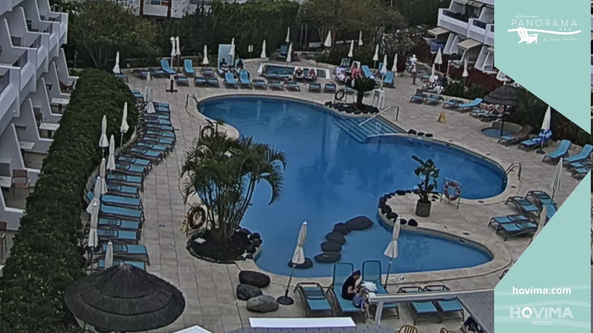 An outdoor pool area with a kidney-shaped pool, numerous lounge chairs, and umbrellas under an overcast sky.