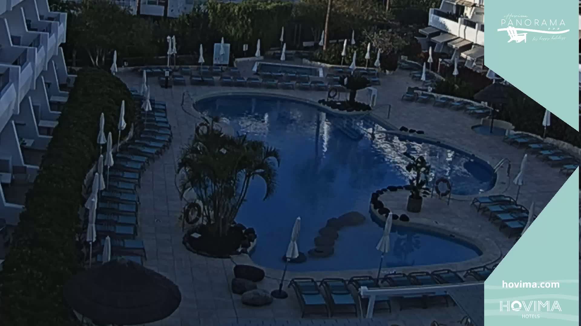 An aerial view shows a resort pool area with numerous lounge chairs and umbrellas surrounding a large, dark blue swimming pool under a dimly lit sky.