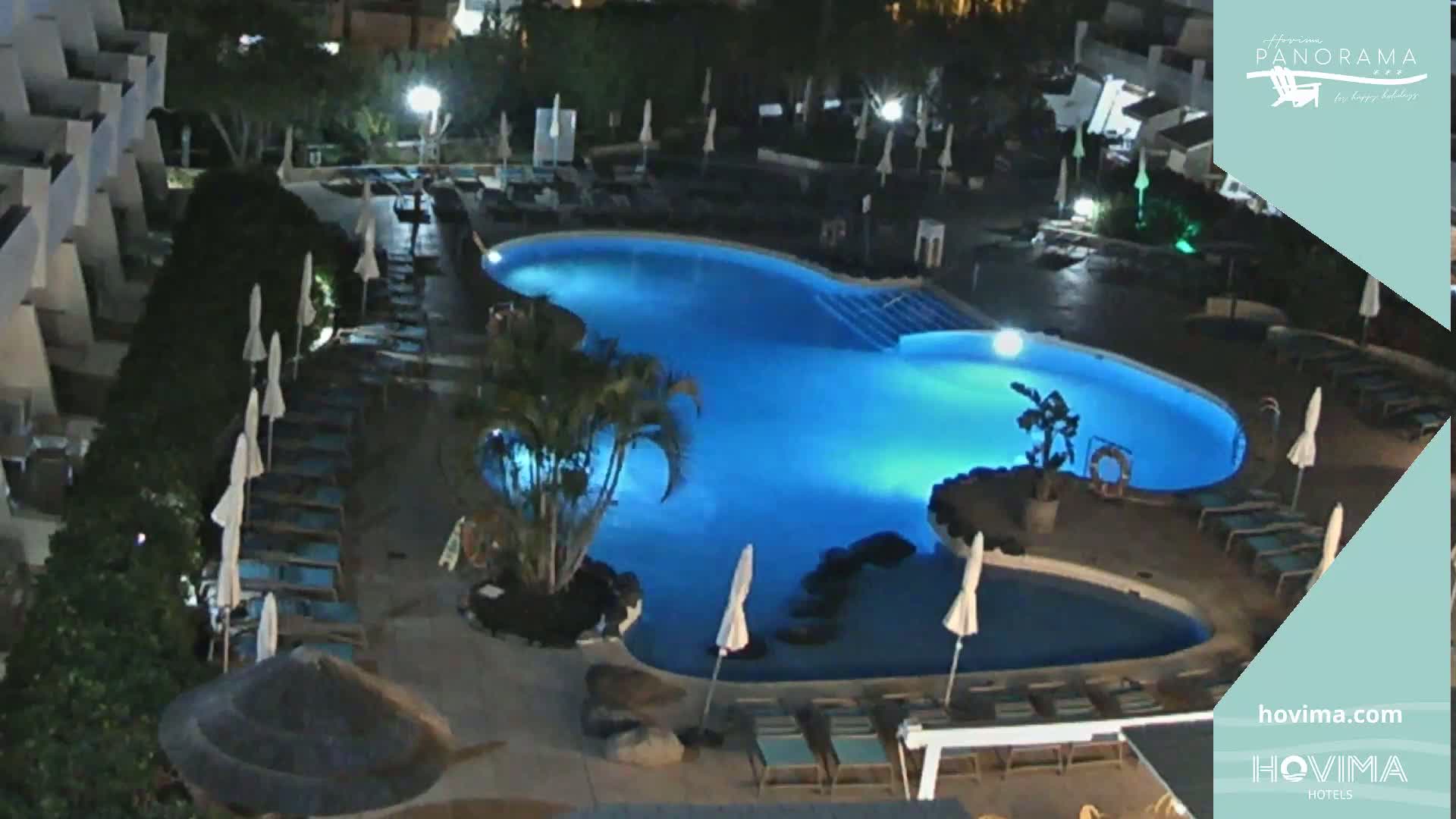 An illuminated swimming pool at night, surrounded by lounge chairs and umbrellas, sits beside a building in a landscaped area.