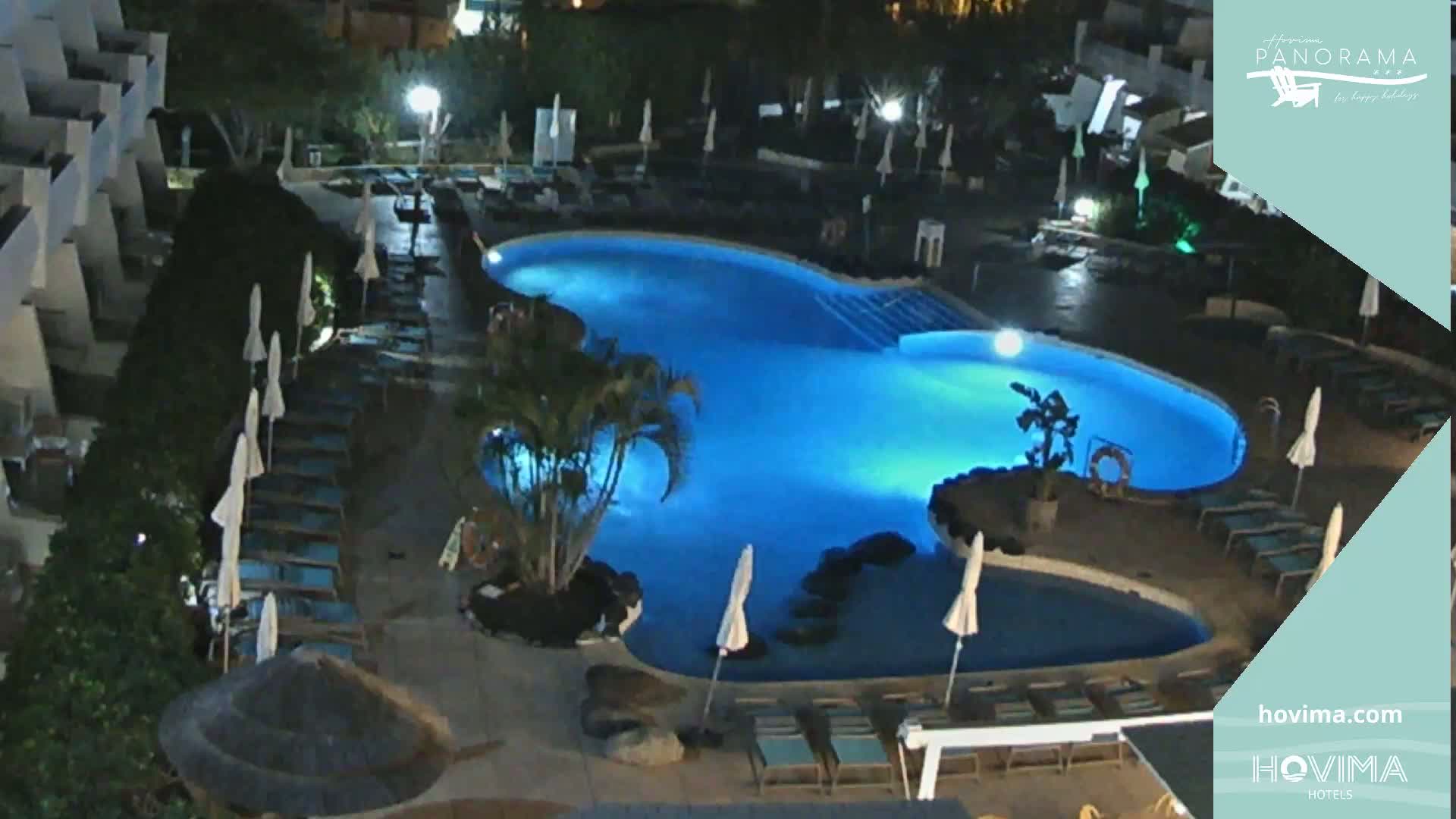 An illuminated, kidney-shaped swimming pool surrounded by lounge chairs and umbrellas at night.