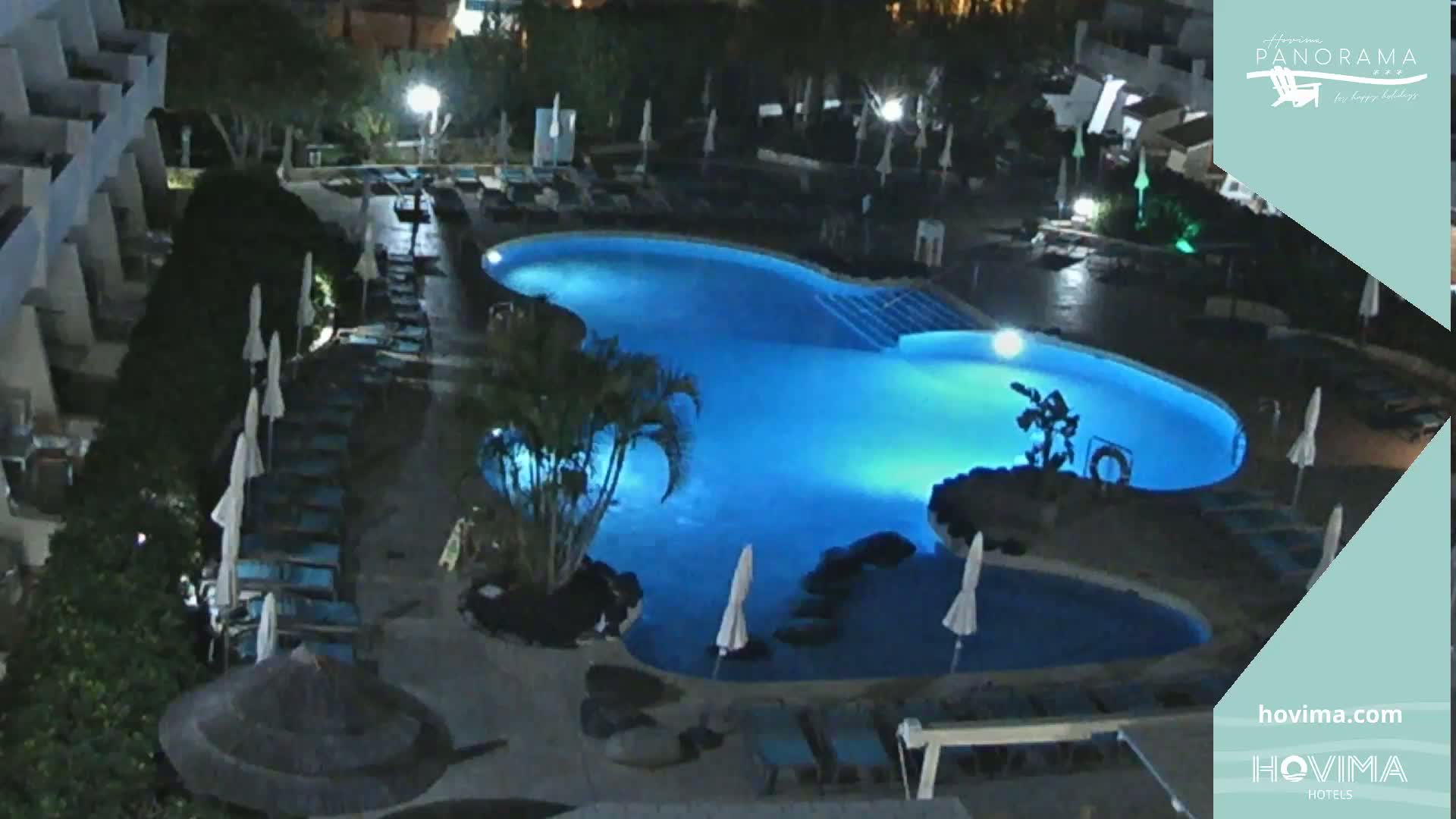 An illuminated, kidney-shaped swimming pool is surrounded by lounge chairs and umbrellas at night.
