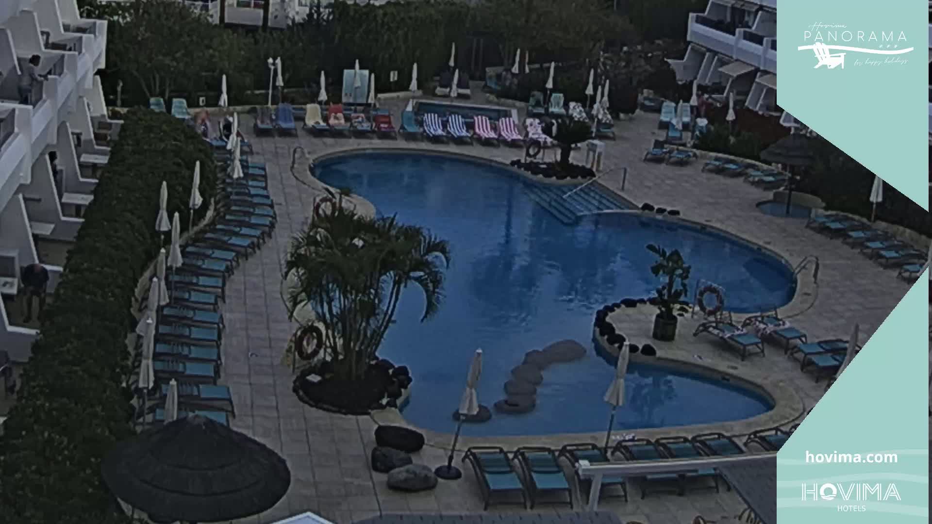 An overhead view shows a kidney-shaped swimming pool surrounded by lounge chairs and umbrellas at a resort, under an overcast sky.