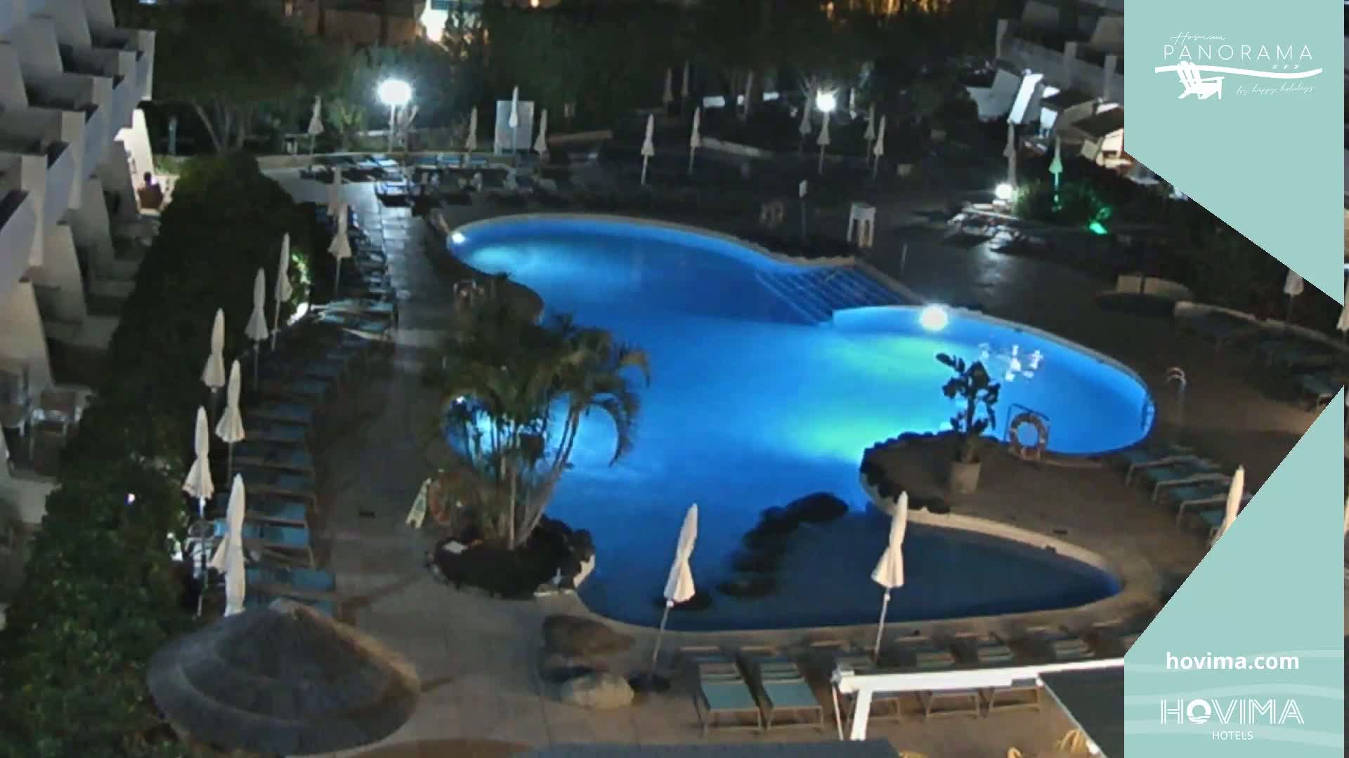 A nighttime view of a resort pool area with surrounding lounge chairs and palm trees.