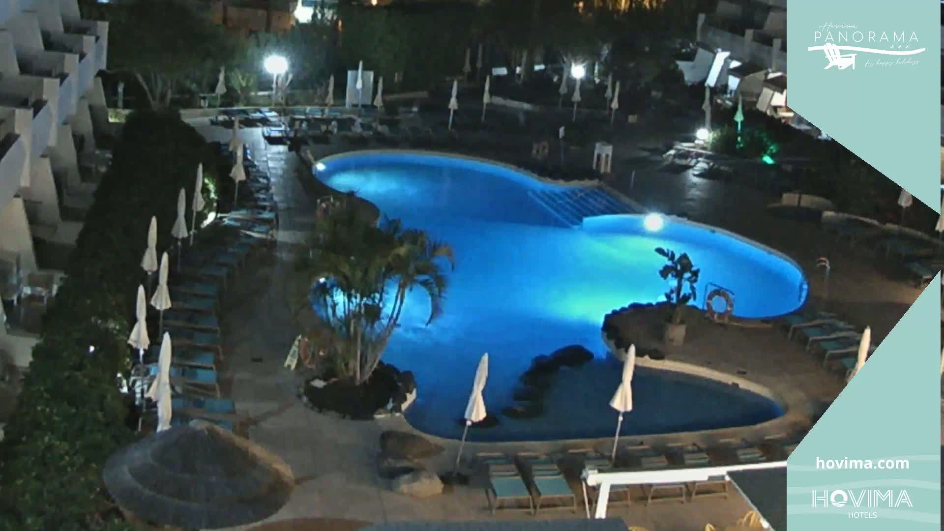 An illuminated swimming pool at night, surrounded by lounge chairs and umbrellas, next to a building.