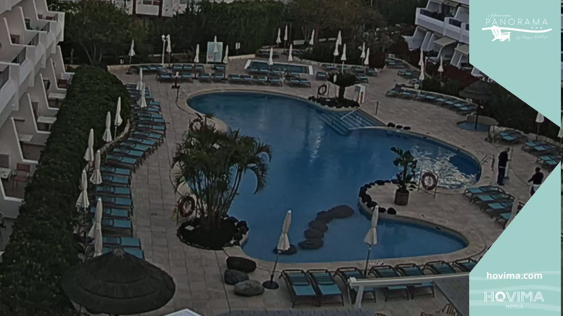 An outdoor pool with numerous lounge chairs and umbrellas surrounding it, situated beside a building on a mostly overcast day.