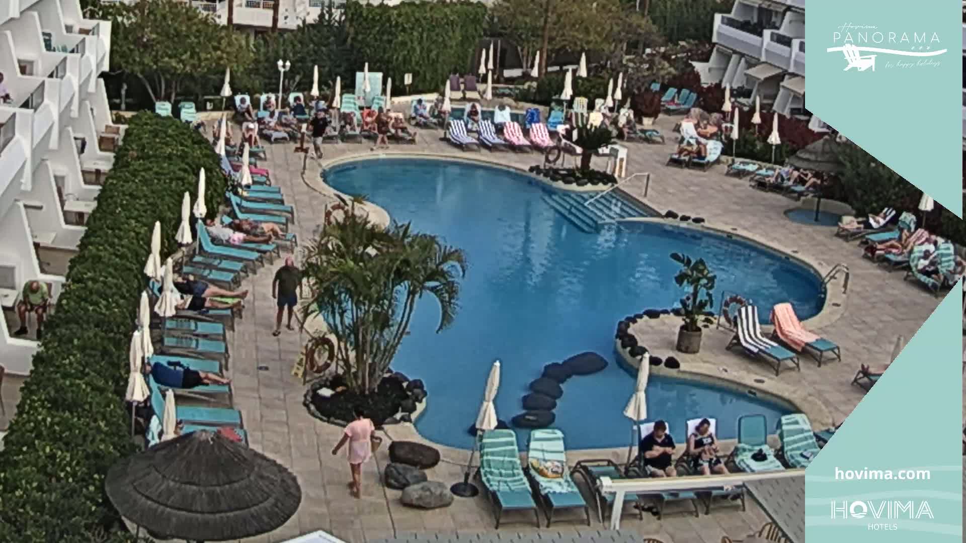 An outdoor pool area with many people lounging on chairs and sunbathing around a kidney-shaped swimming pool.