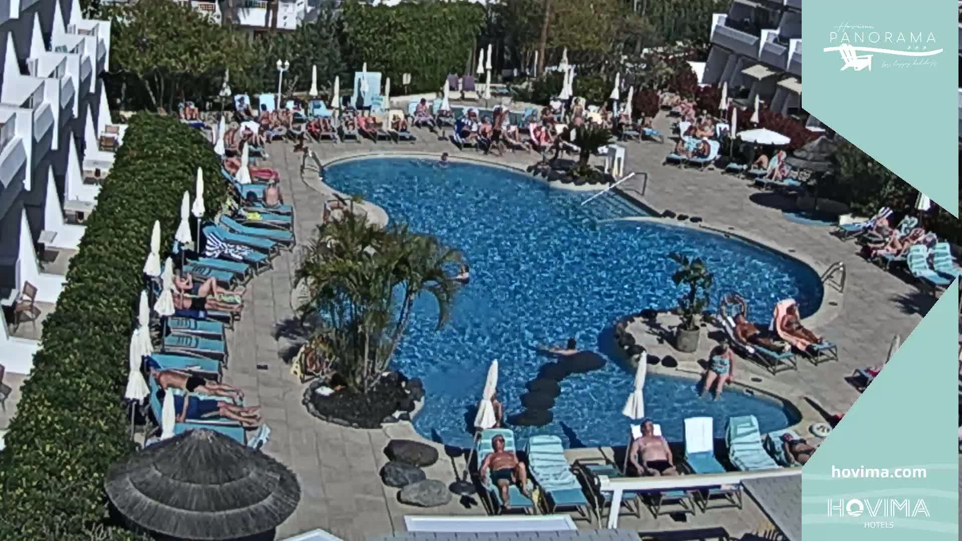 A sunny outdoor scene shows a resort pool surrounded by lounge chairs and umbrellas, with numerous people swimming and sunbathing.