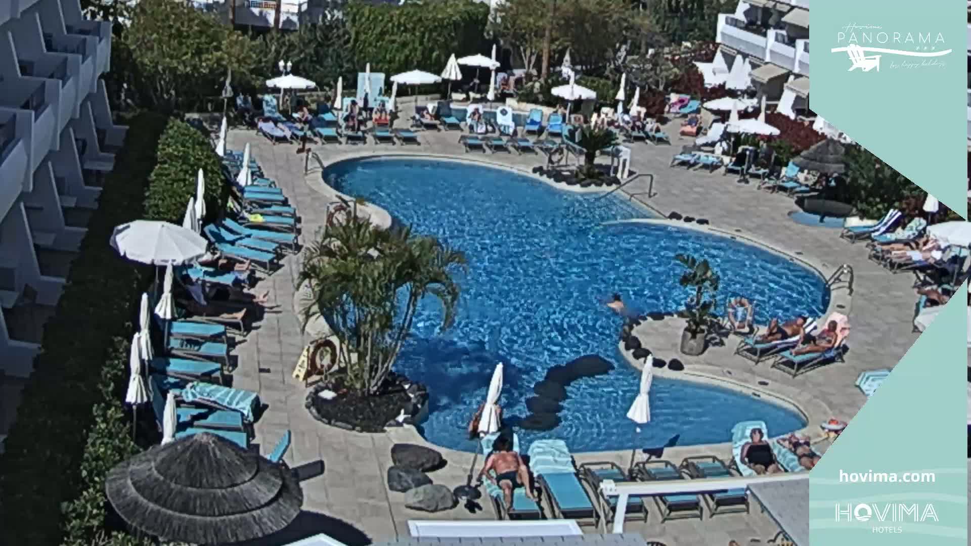 An outdoor pool area with many people lounging on chairs and in the water on a sunny day.