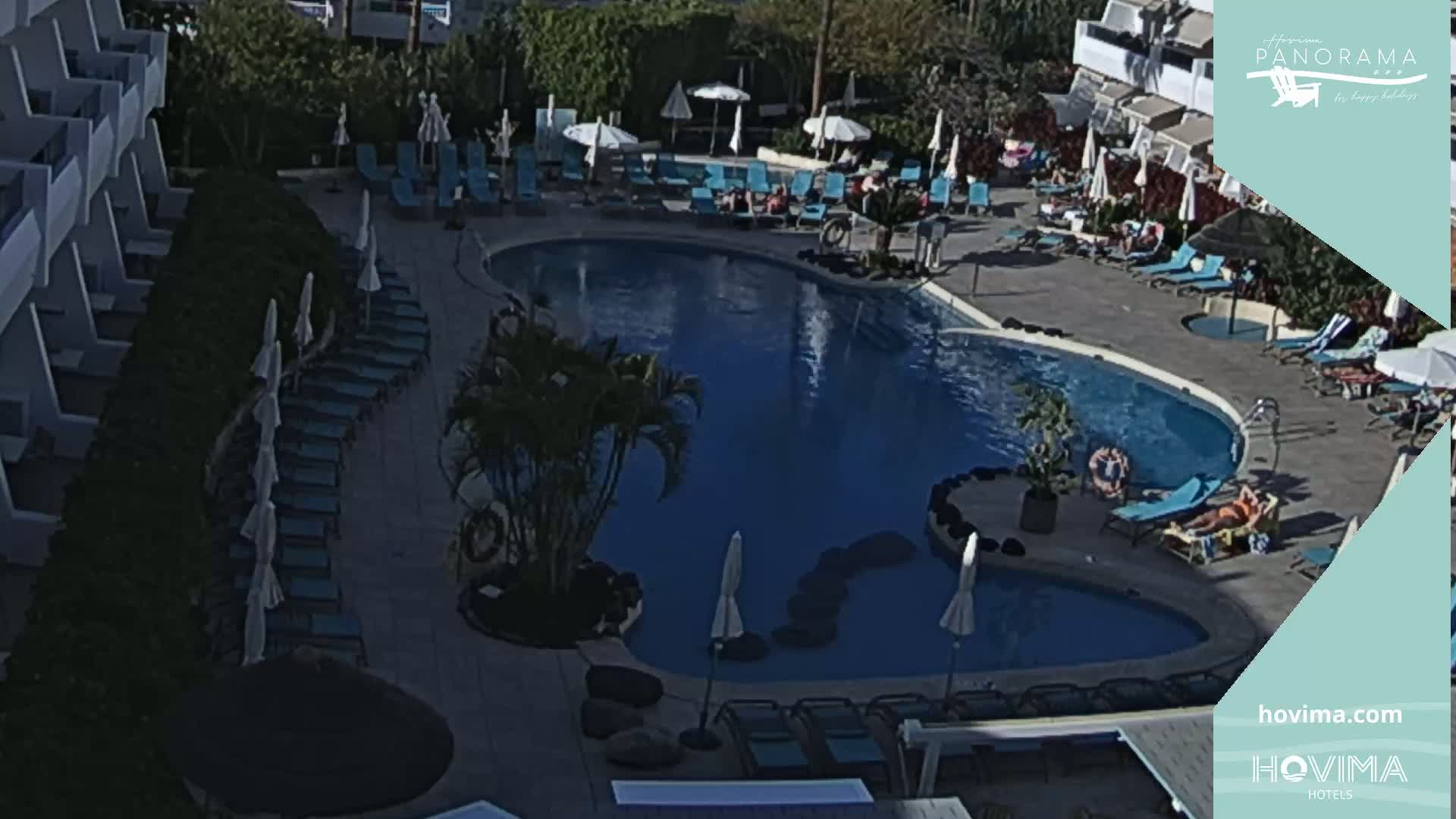 An outdoor pool area with numerous lounge chairs and umbrellas surrounding a kidney-shaped pool, under sunny conditions.
