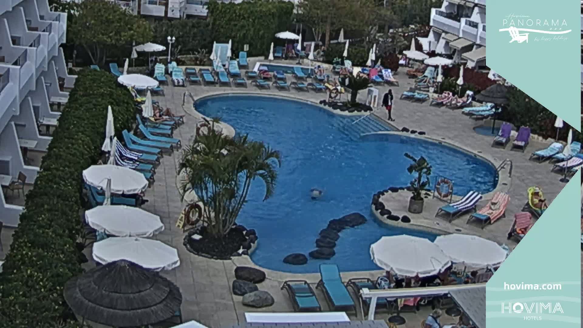 An outdoor swimming pool surrounded by lounge chairs, umbrellas, and a hotel building on a partly cloudy day.