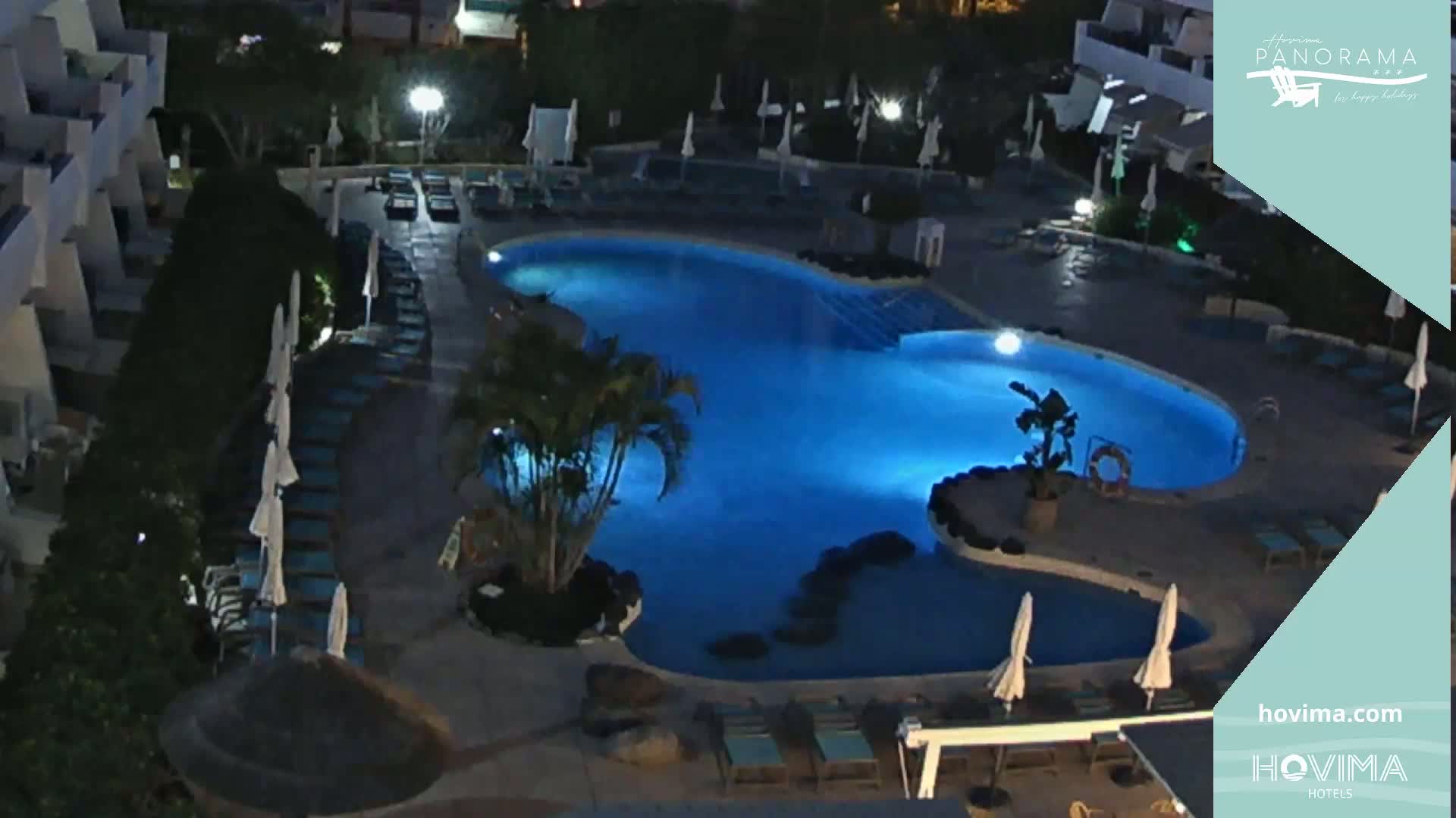 An illuminated, irregularly shaped swimming pool at night, surrounded by lounge chairs and umbrellas, is nestled within a resort complex.