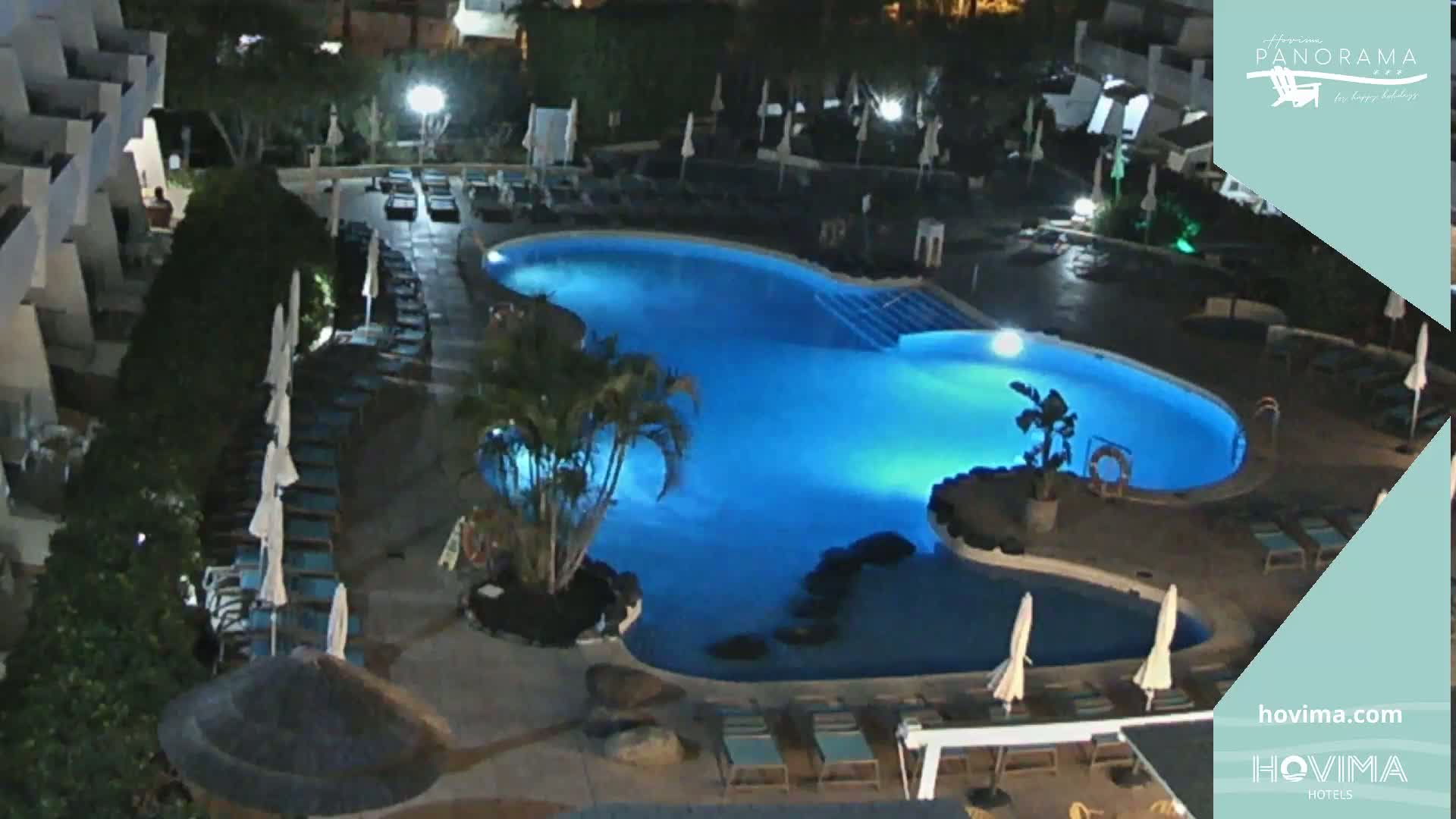 An illuminated swimming pool at night, surrounded by lounge chairs and palm trees, is situated within a resort complex.