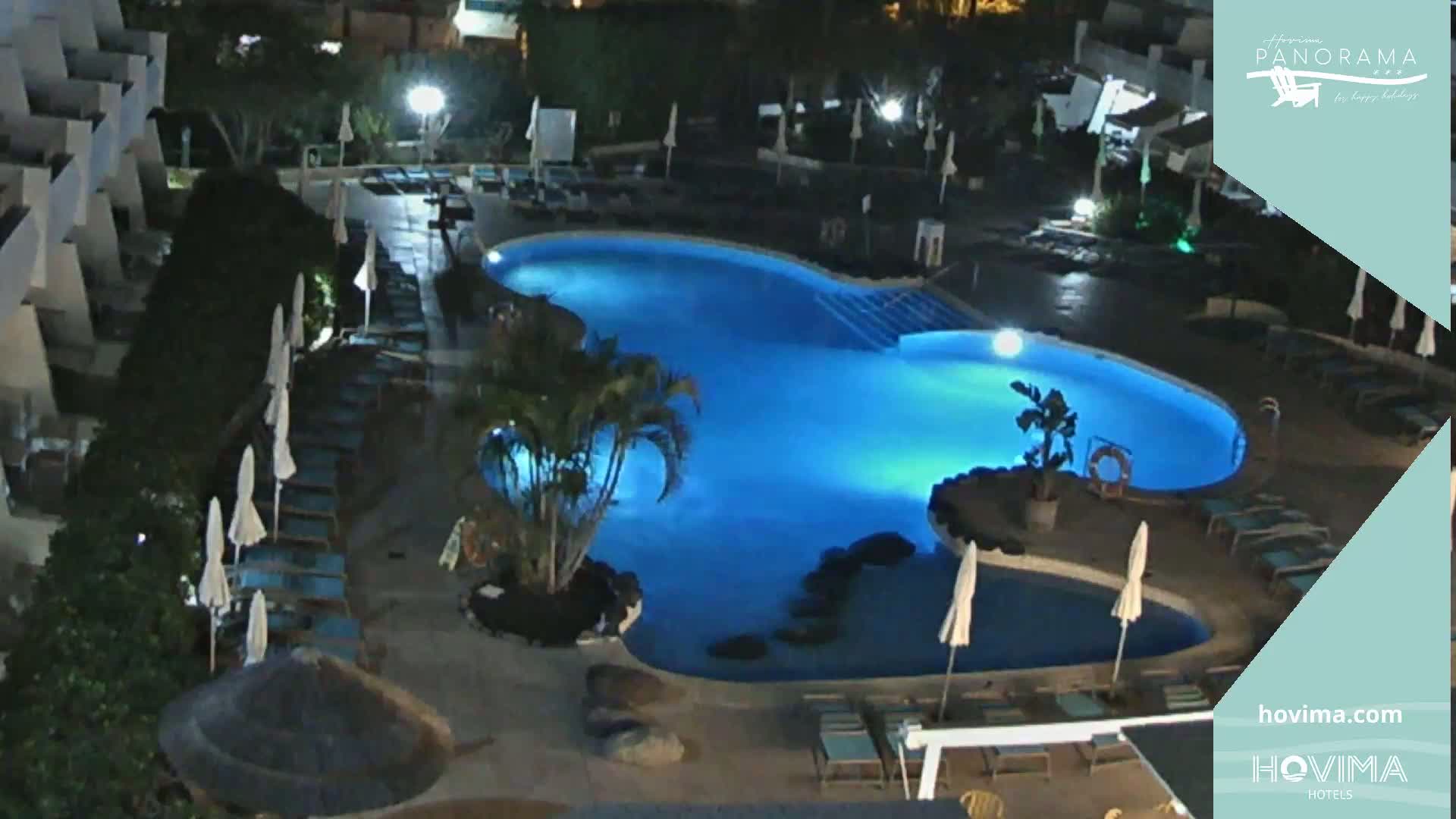 An illuminated swimming pool at night, surrounded by lounge chairs and umbrellas, is situated next to a building.
