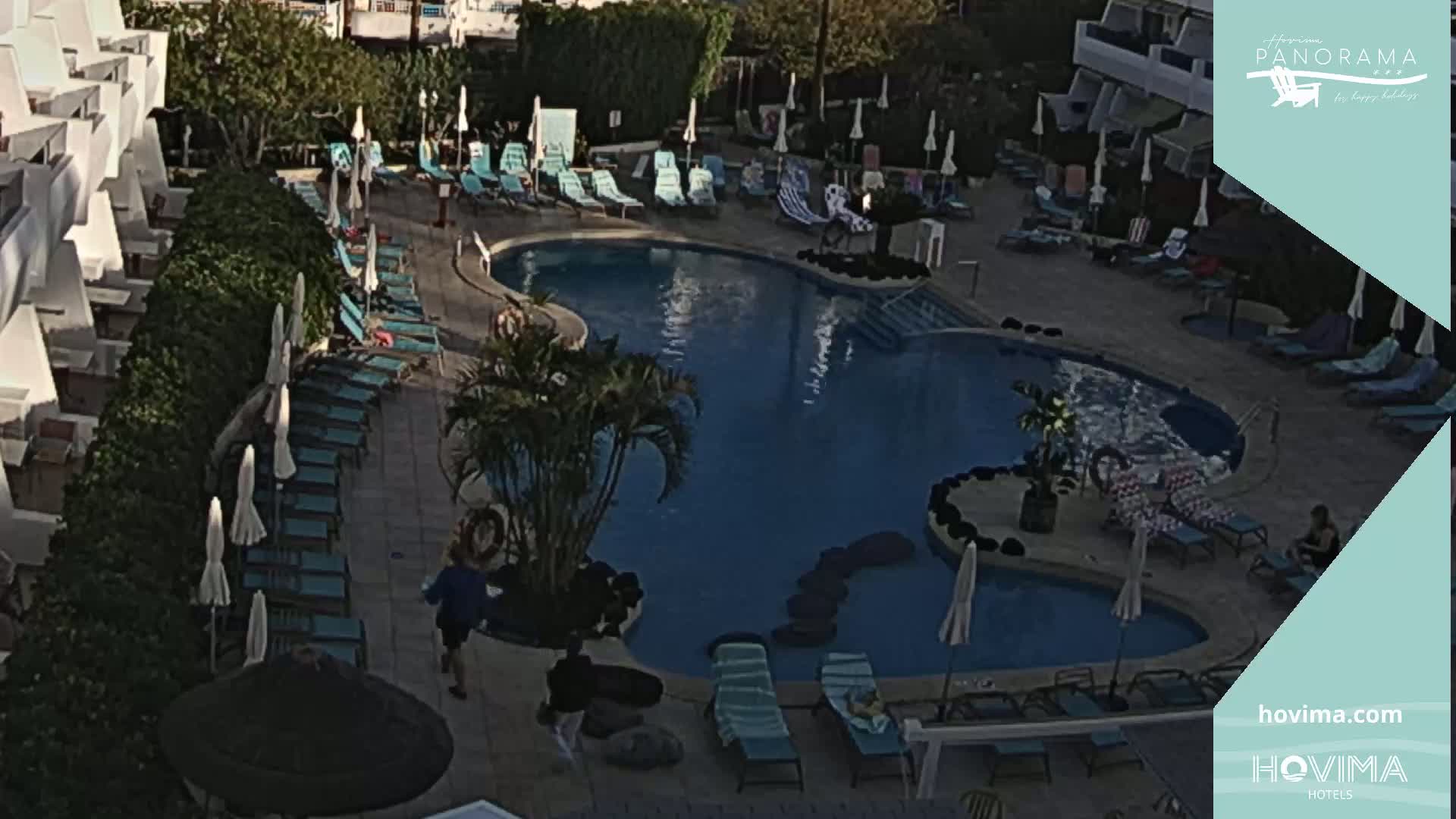 An outdoor swimming pool surrounded by lounge chairs and umbrellas next to a building, under a mostly clear sky.