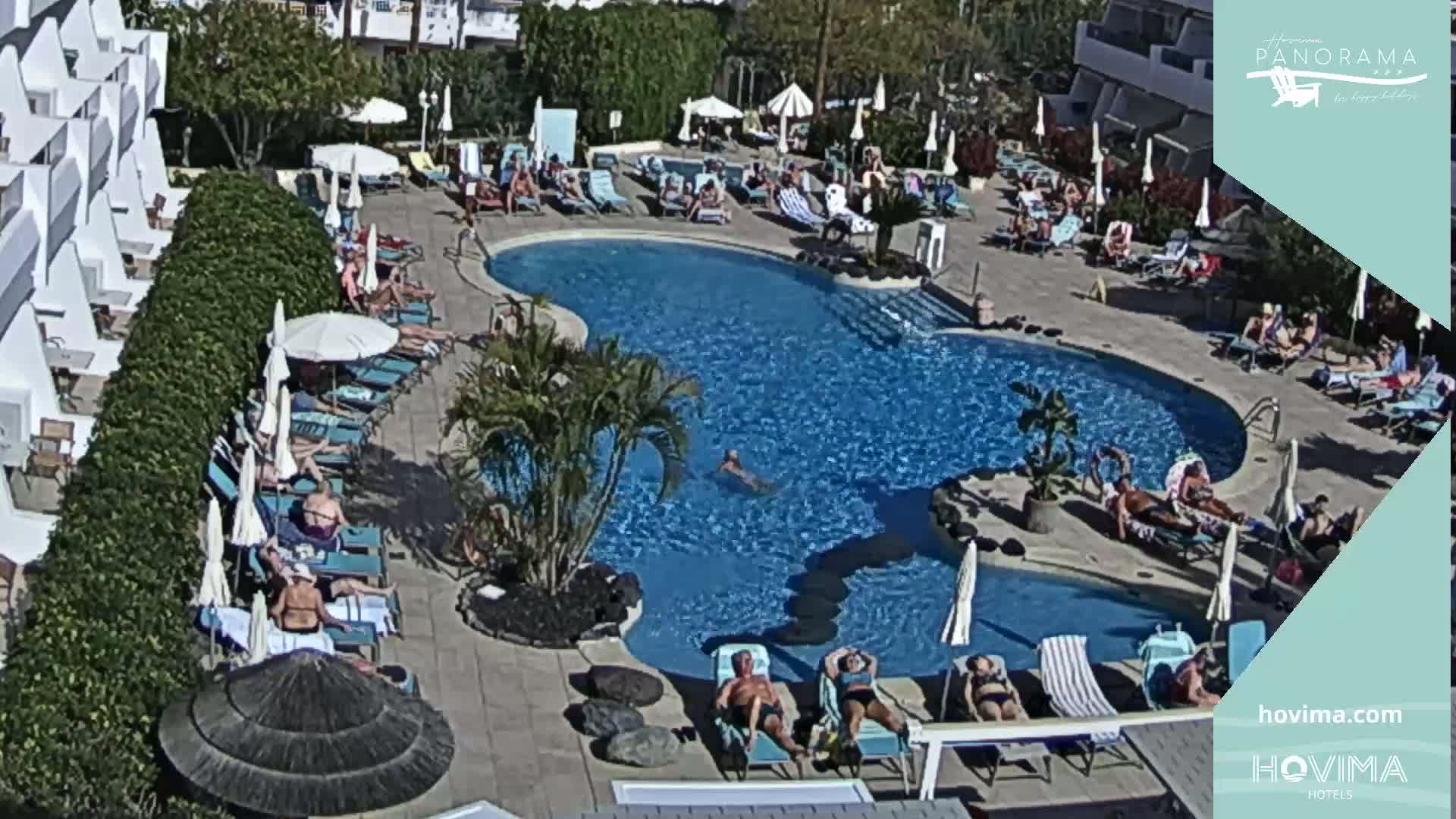 A sunny outdoor scene shows a large swimming pool surrounded by sunbathers lounging on chairs and under umbrellas near a hotel building.