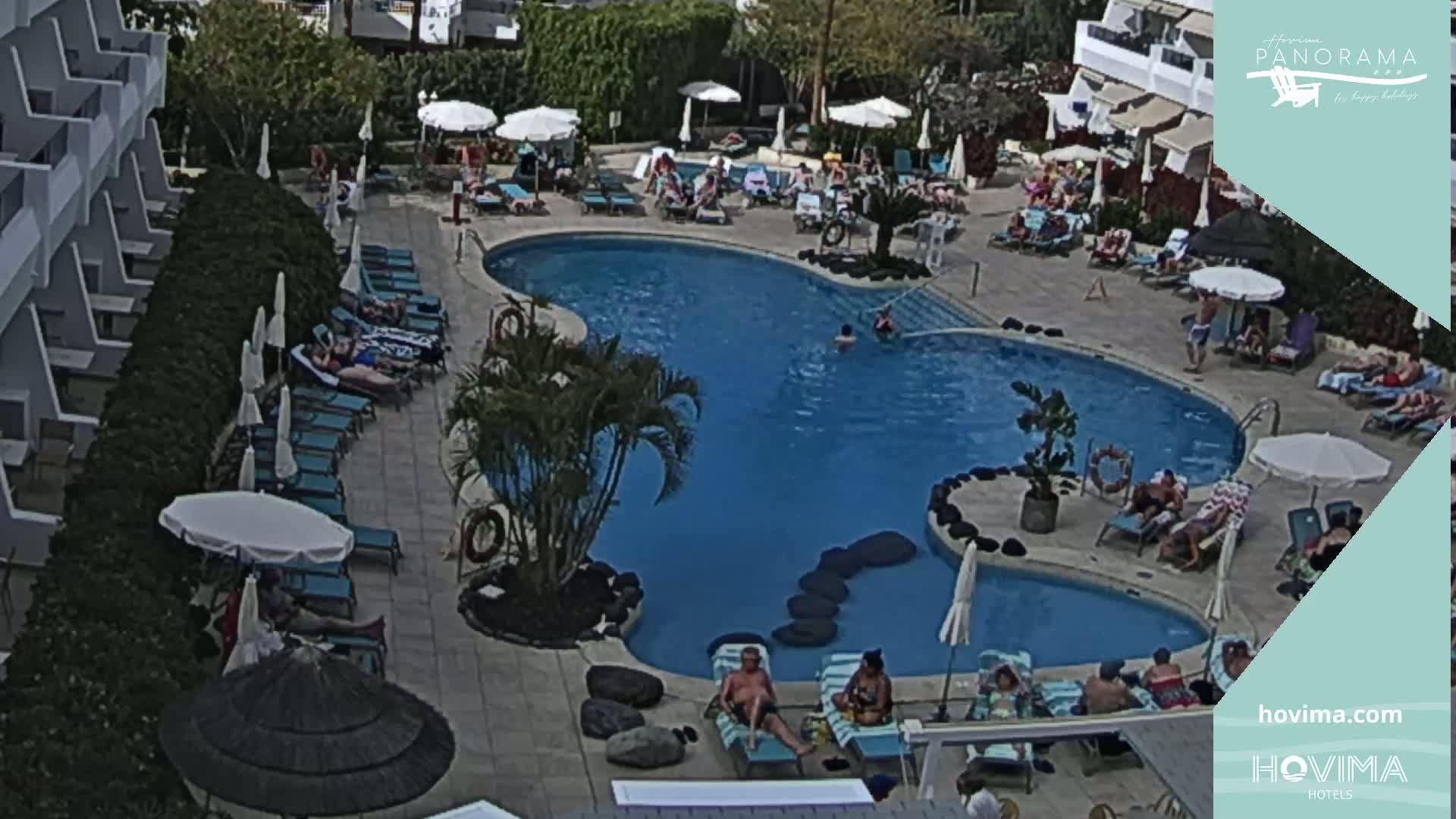 An outdoor swimming pool surrounded by lounge chairs and umbrellas, with many people relaxing and swimming on a sunny day.