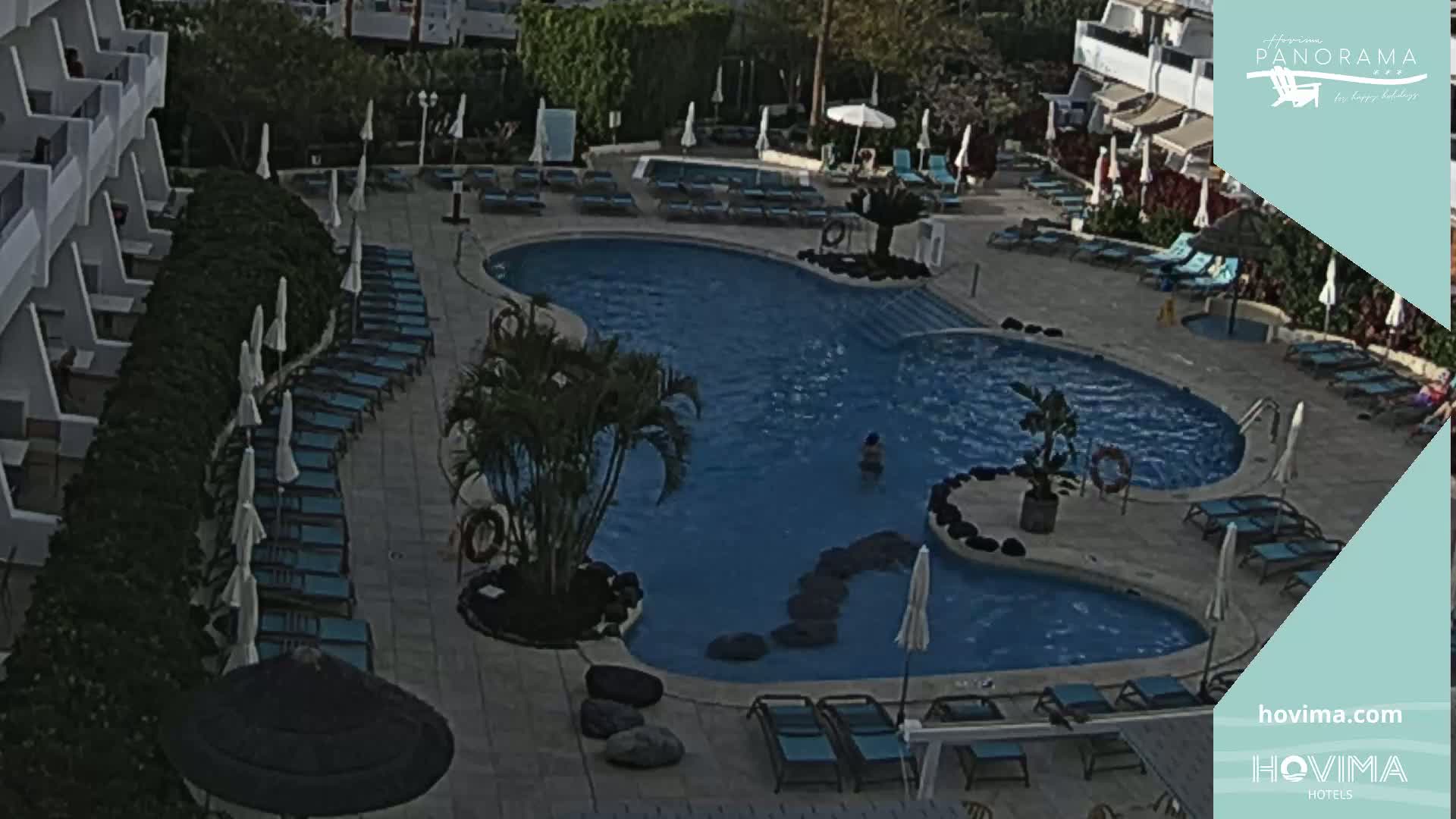 An outdoor pool with surrounding lounge chairs and umbrellas is situated next to a building under a sunny sky.