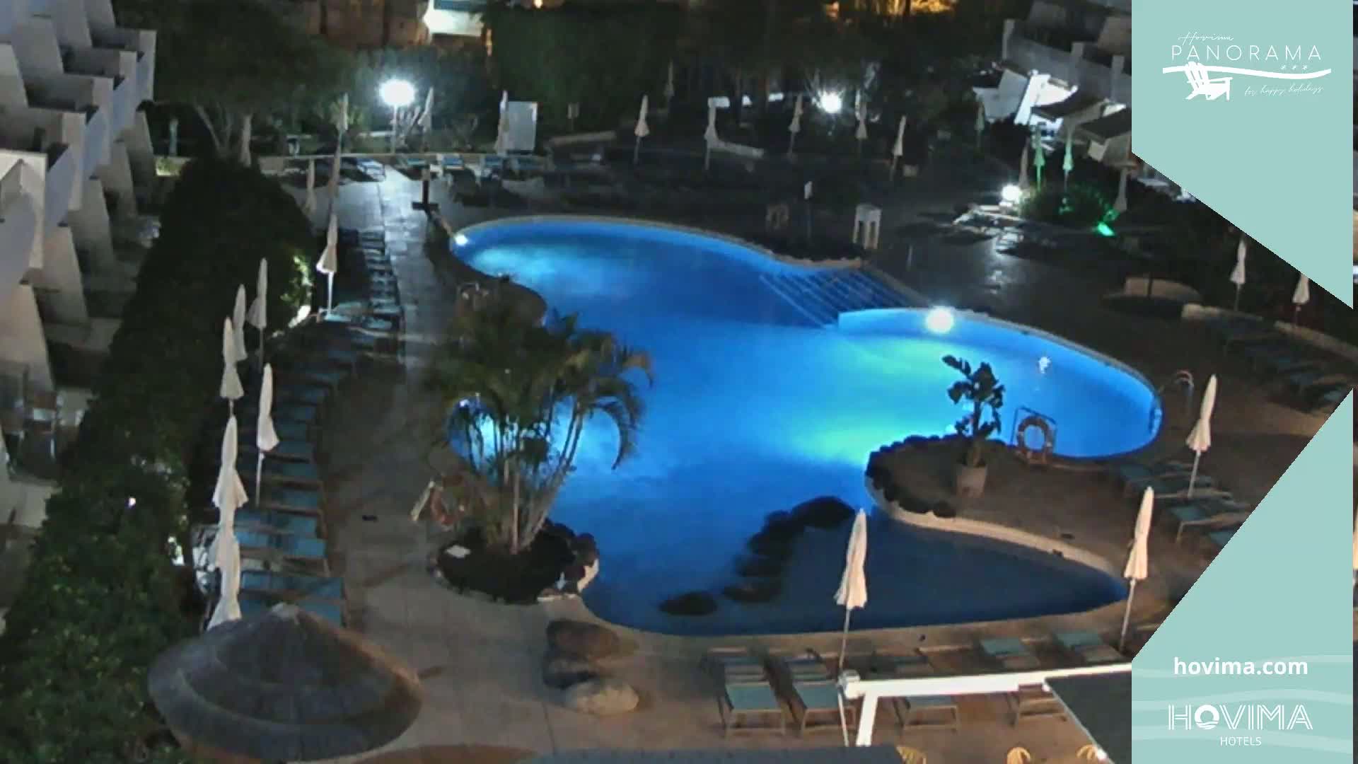 An illuminated, kidney-shaped swimming pool at night, surrounded by lounge chairs and palm trees, is seen from an elevated perspective.