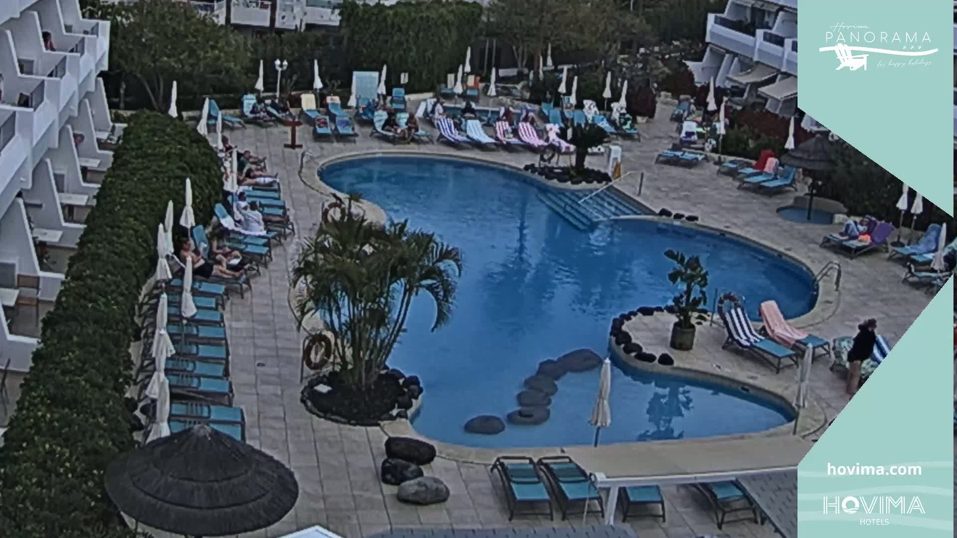 An outdoor pool area with several people lounging on chairs surrounding a kidney-shaped pool, under a partly cloudy sky.