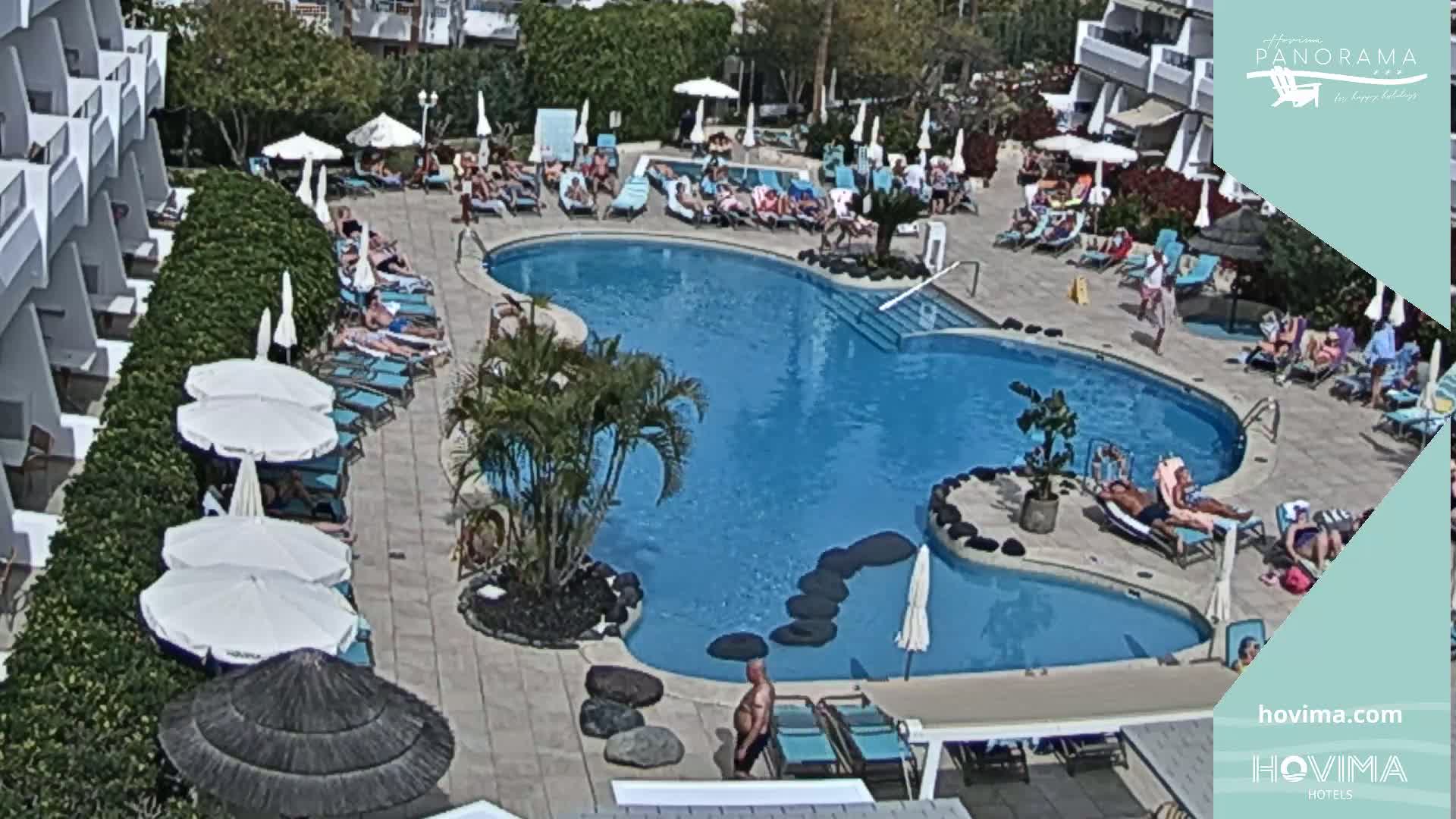 An outdoor pool area with many people relaxing on lounge chairs and in the water on a sunny day.