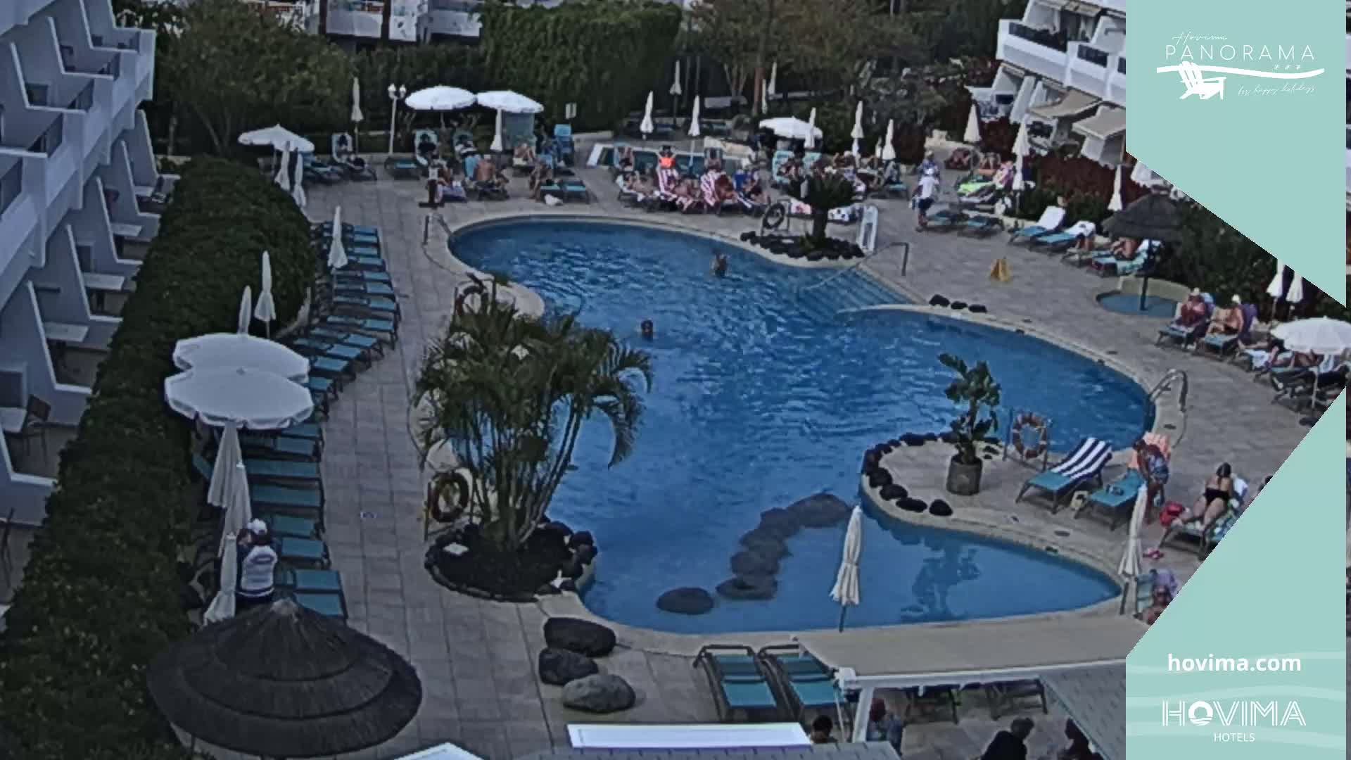 An outdoor swimming pool surrounded by lounge chairs, umbrellas, and a building, with several people relaxing by the pool.
