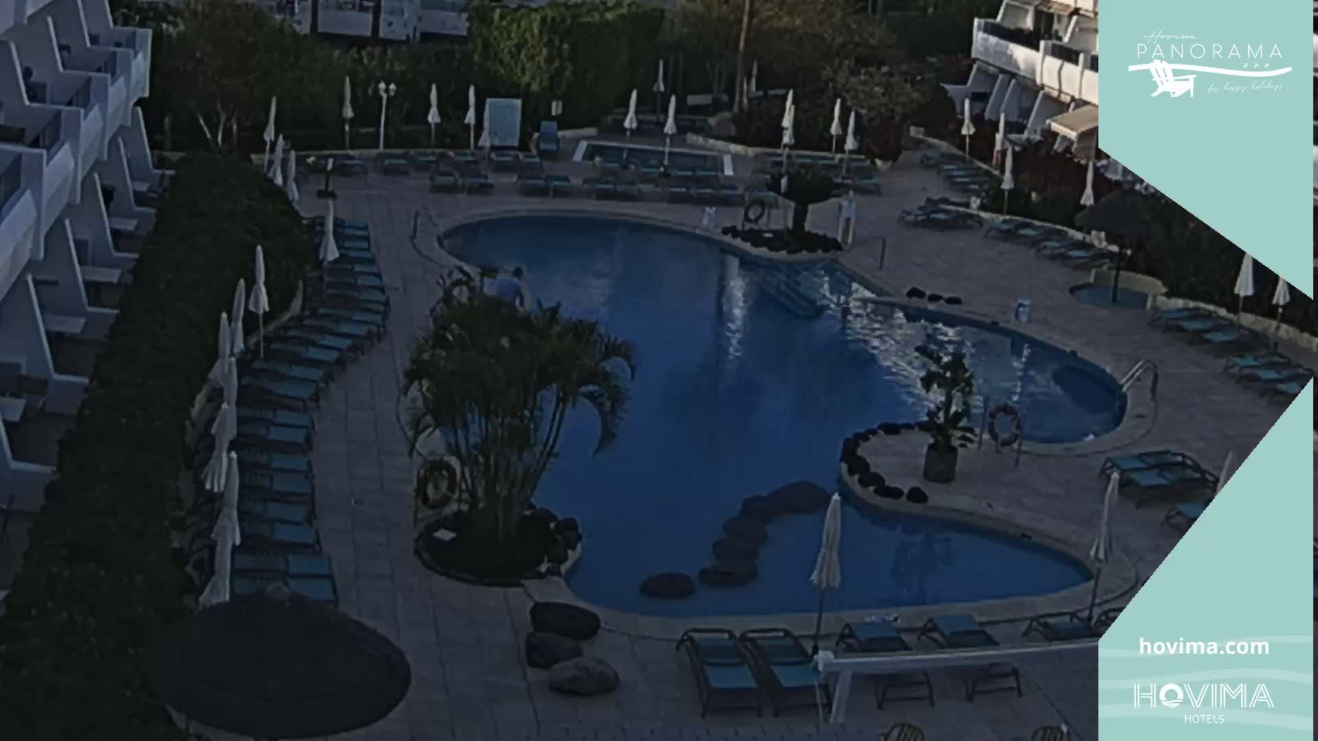 An aerial view shows a kidney-shaped swimming pool surrounded by lounge chairs and umbrellas at a resort.