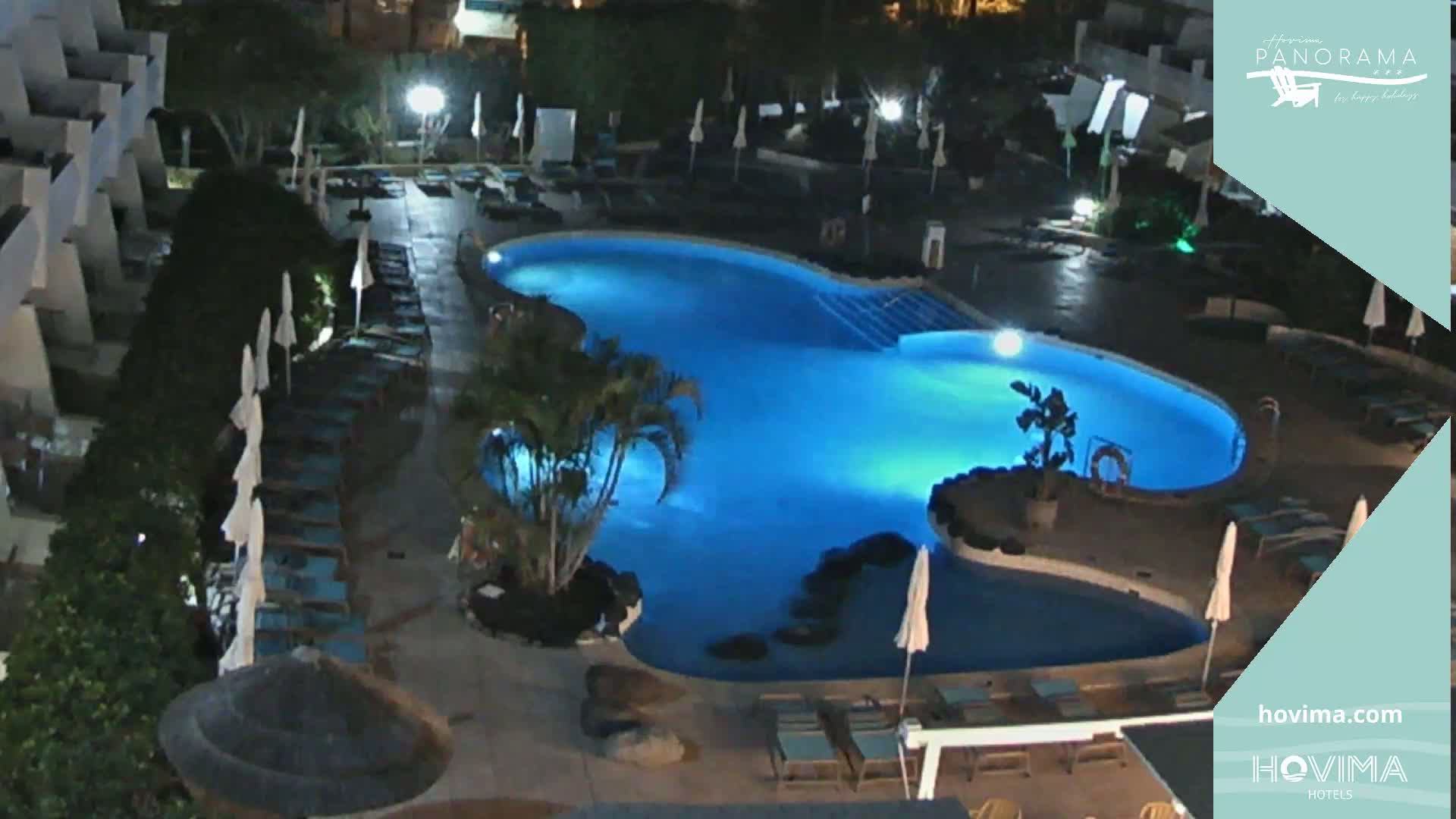 An illuminated, kidney-shaped swimming pool surrounded by lounge chairs and umbrellas at night.