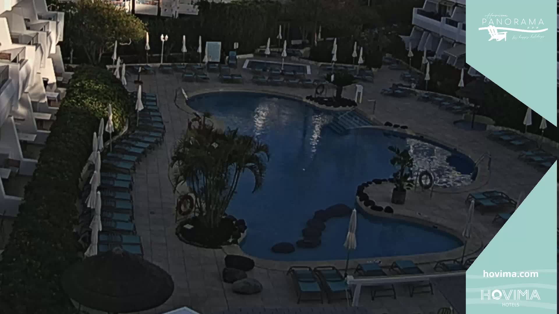 An outdoor pool with surrounding lounge chairs and umbrellas sits next to a building in the evening.