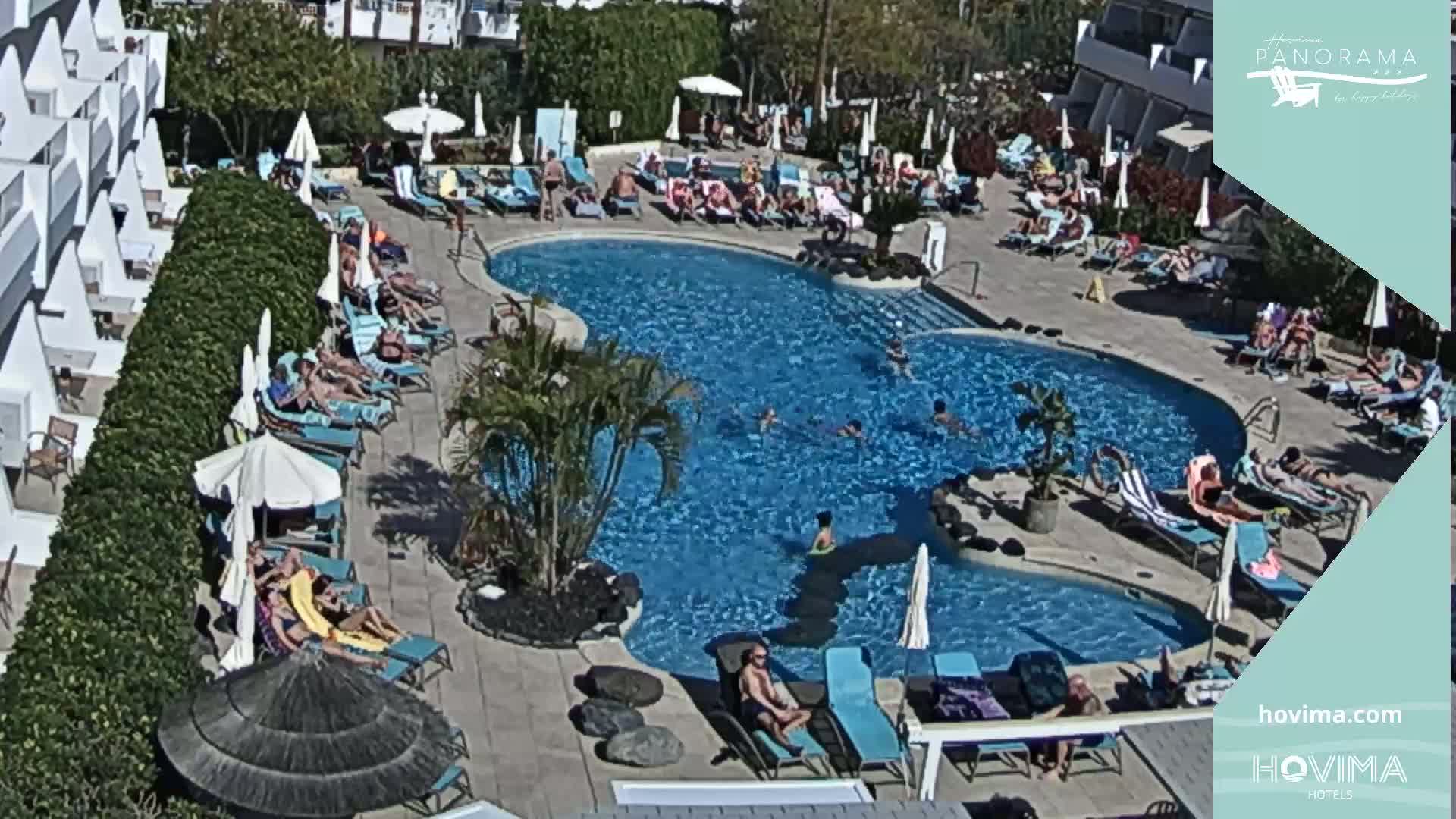 A large outdoor swimming pool surrounded by sunbathers relaxing on lounge chairs under umbrellas on a sunny day.