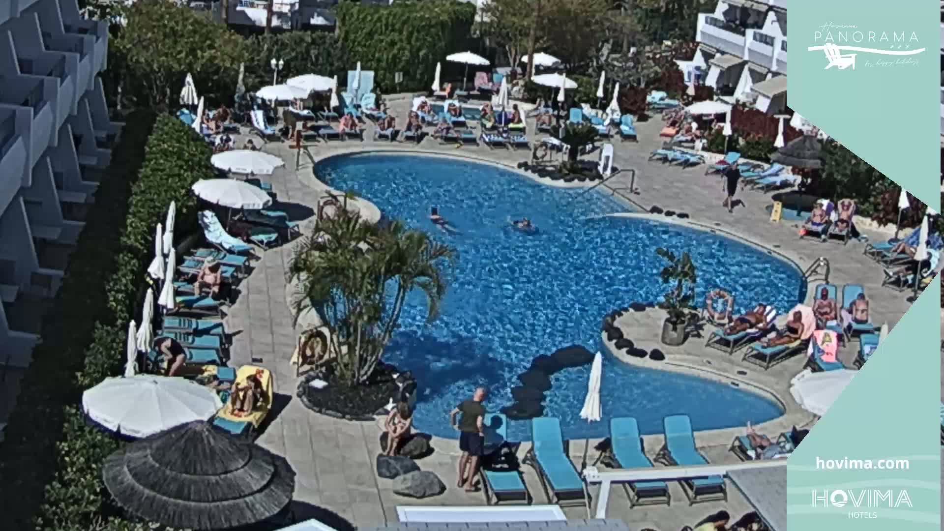 An outdoor pool area with many people lounging on chairs and in the water under a sunny sky.