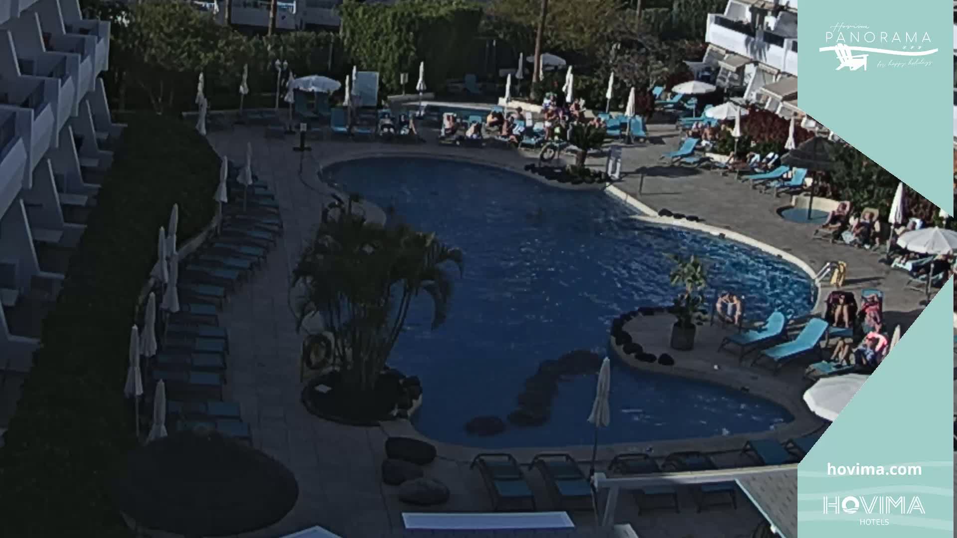 An outdoor pool with sunbathers and lounge chairs is surrounded by a building and landscaping under sunny conditions.