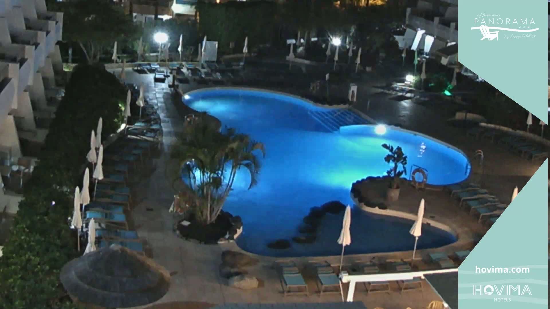 A brightly lit swimming pool at night, surrounded by lounge chairs and palm trees.