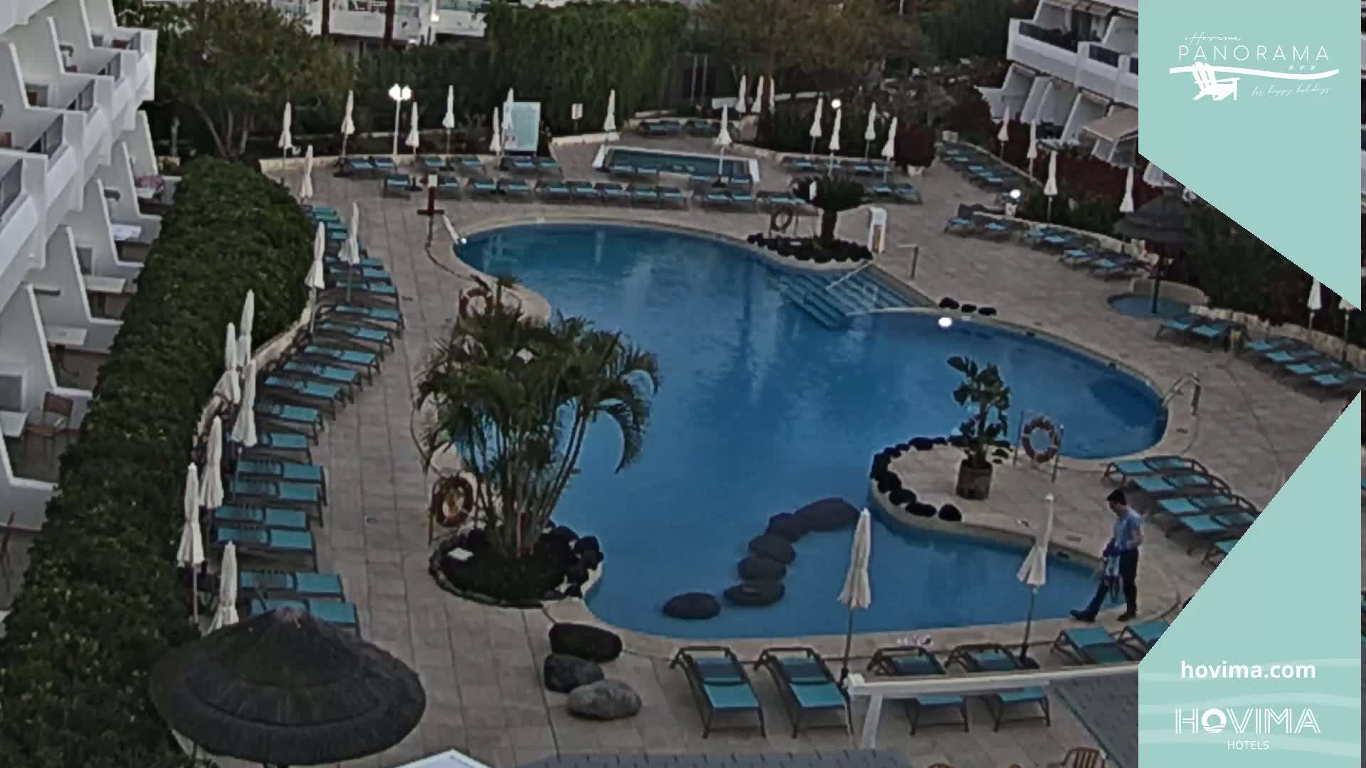 An outdoor pool area with numerous lounge chairs and umbrellas surrounding a kidney-shaped pool, situated next to a building.