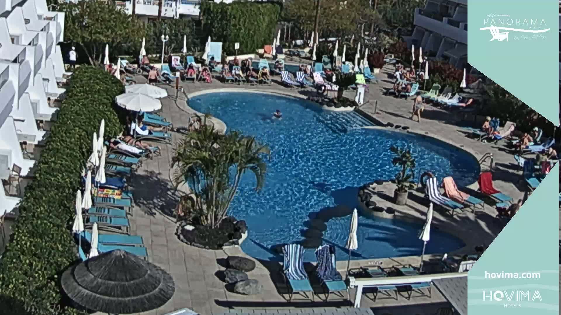 A sunny outdoor swimming pool area with numerous lounge chairs and umbrellas surrounding it, next to a white building.