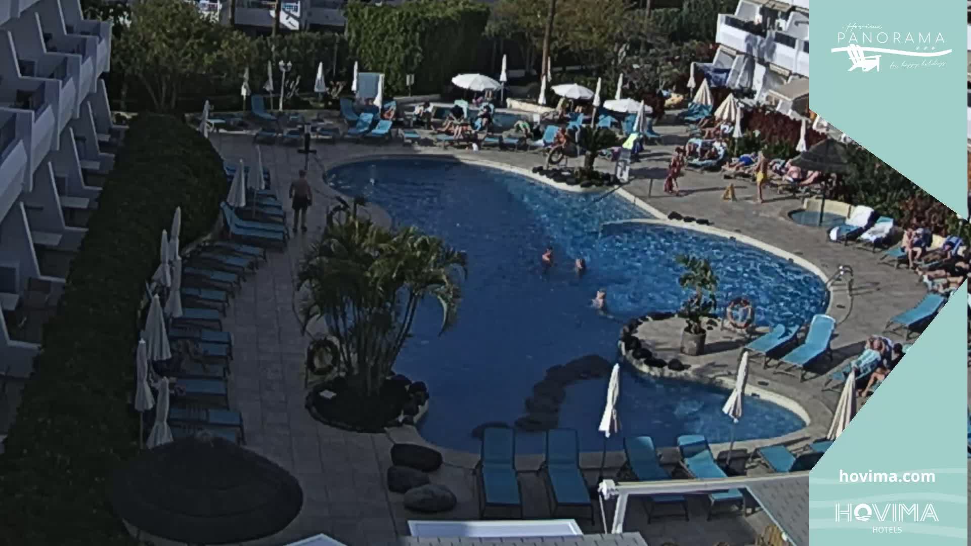 A sunny outdoor scene shows a kidney-shaped swimming pool surrounded by lounge chairs and umbrellas, with several people swimming and relaxing.