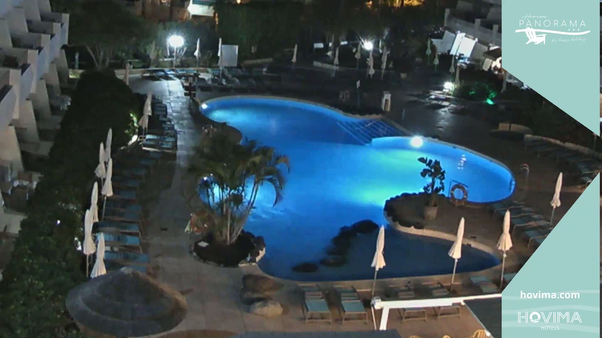 An illuminated outdoor swimming pool at night, surrounded by lounge chairs and umbrellas, next to a building.