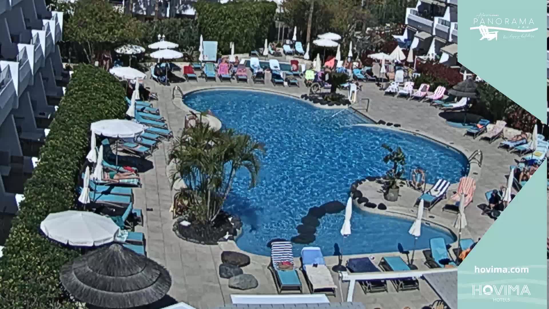 A sunny outdoor scene shows a kidney-shaped swimming pool surrounded by lounge chairs, umbrellas, and people relaxing.