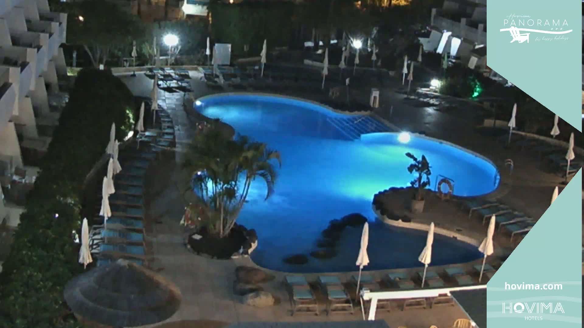 An illuminated swimming pool surrounded by lounge chairs and umbrellas at night.