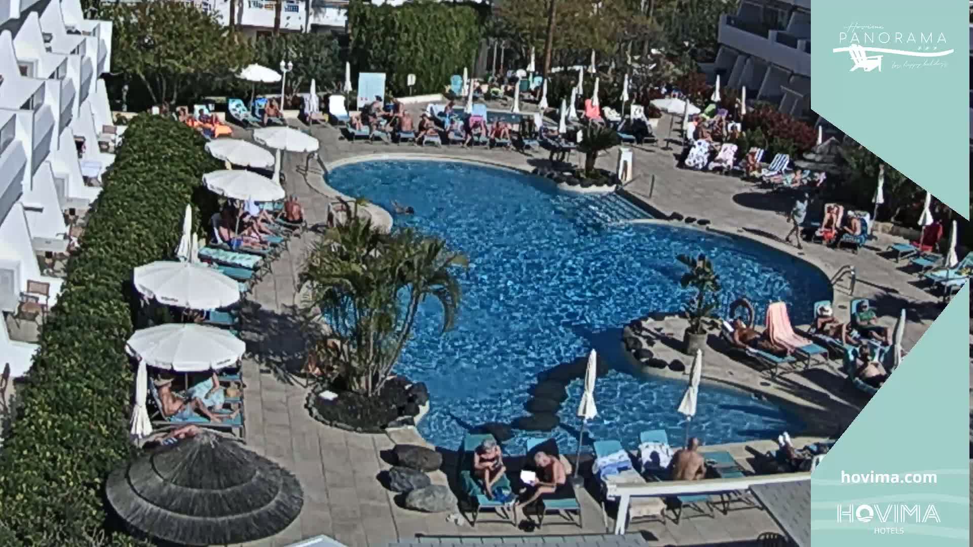 A sunny outdoor pool area with many people lounging on chairs and in the water, surrounded by white umbrellas and palm trees.