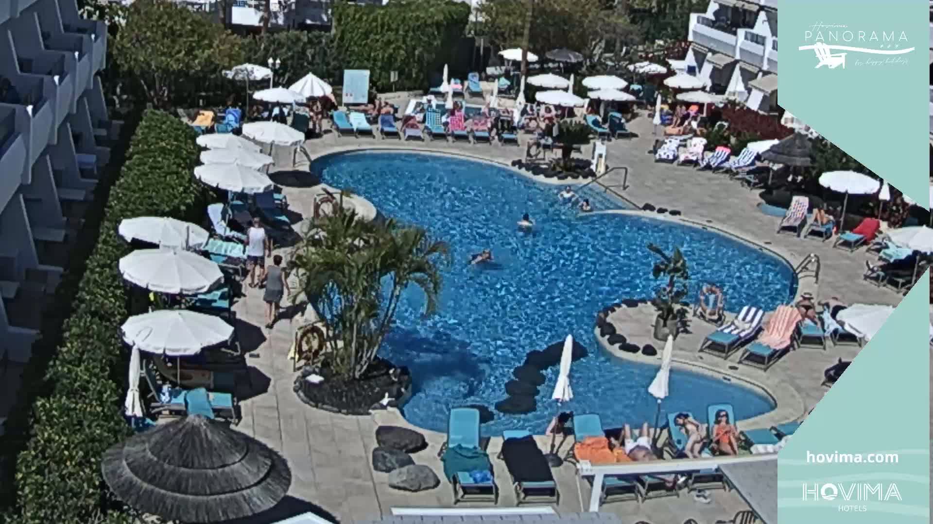 A sunny outdoor pool area with numerous people swimming and lounging on chairs under umbrellas near a building.
