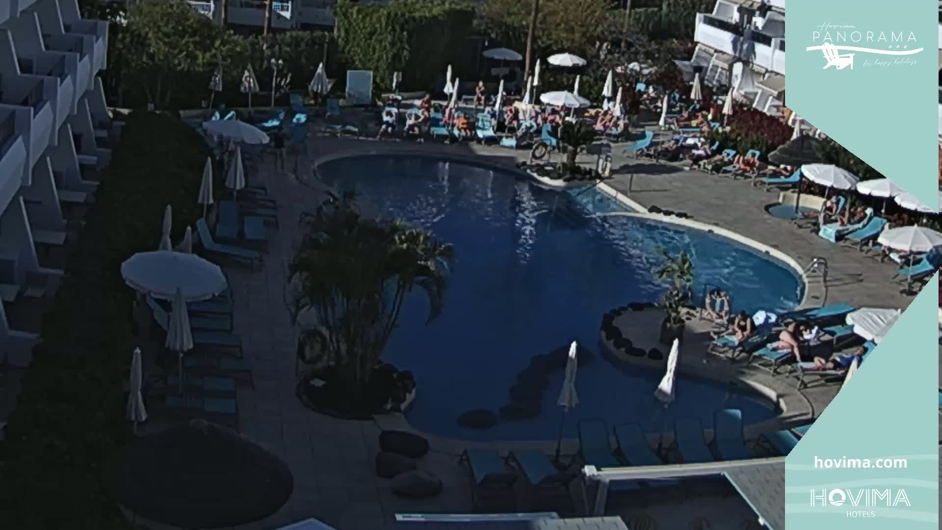 An outdoor pool area with sunbathers and lounge chairs under umbrellas on a sunny day.