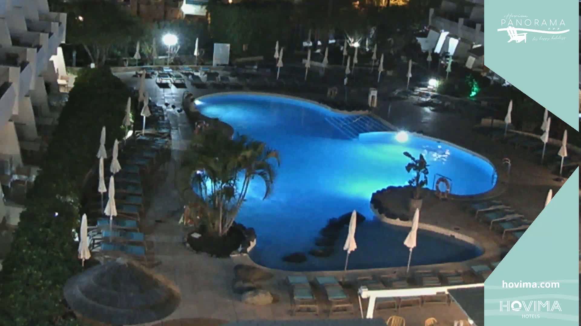 An illuminated kidney-shaped swimming pool at night is surrounded by lounge chairs and umbrellas, situated within a resort complex.