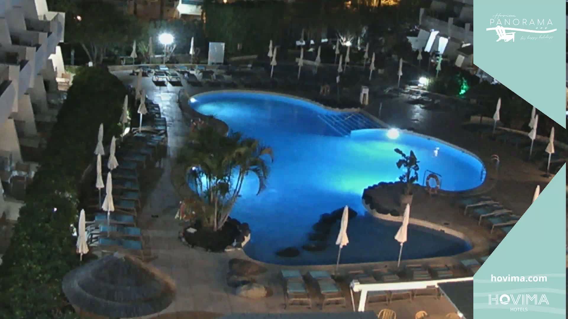 An illuminated swimming pool at night, surrounded by lounge chairs and umbrellas, is situated next to a building.