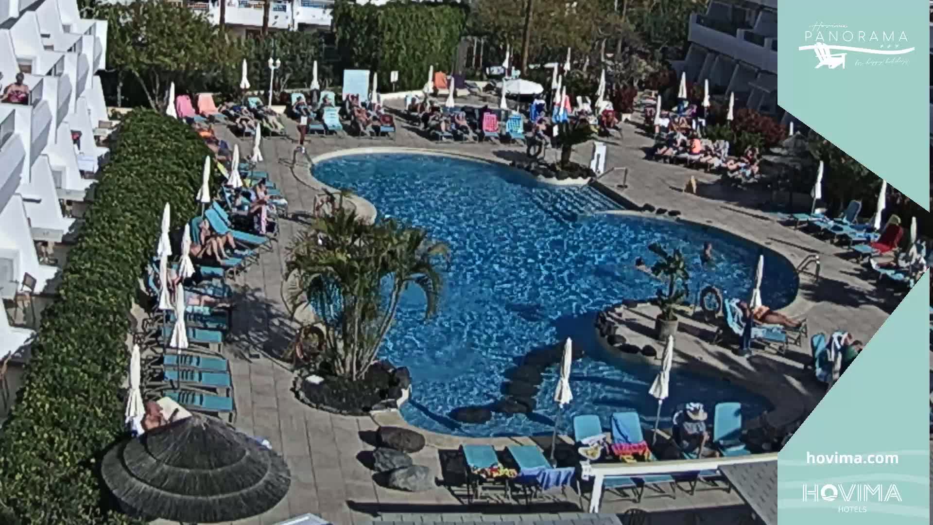 A sunny outdoor scene shows a swimming pool surrounded by lounge chairs and people relaxing on a warm day.